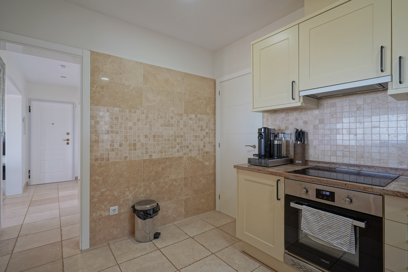 Cuisine fonctionnelle avec placards crème, plan de travail en granit et électroménager. Le mur est revêtu de pierre naturelle avec un détail élégant en mosaïque.