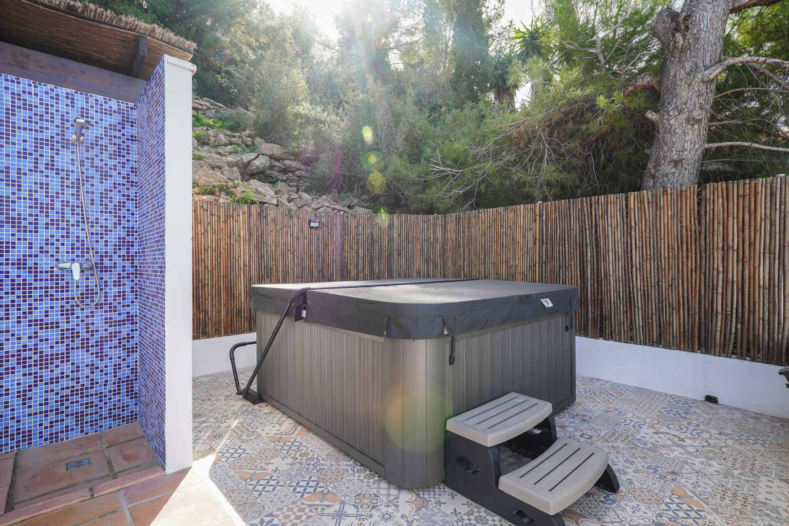 Espace bien-être extérieur avec jacuzzi couvert, douche en mosaïque bleue et sol en carreaux de ciment, entouré d'une clôture en canisse naturelle.