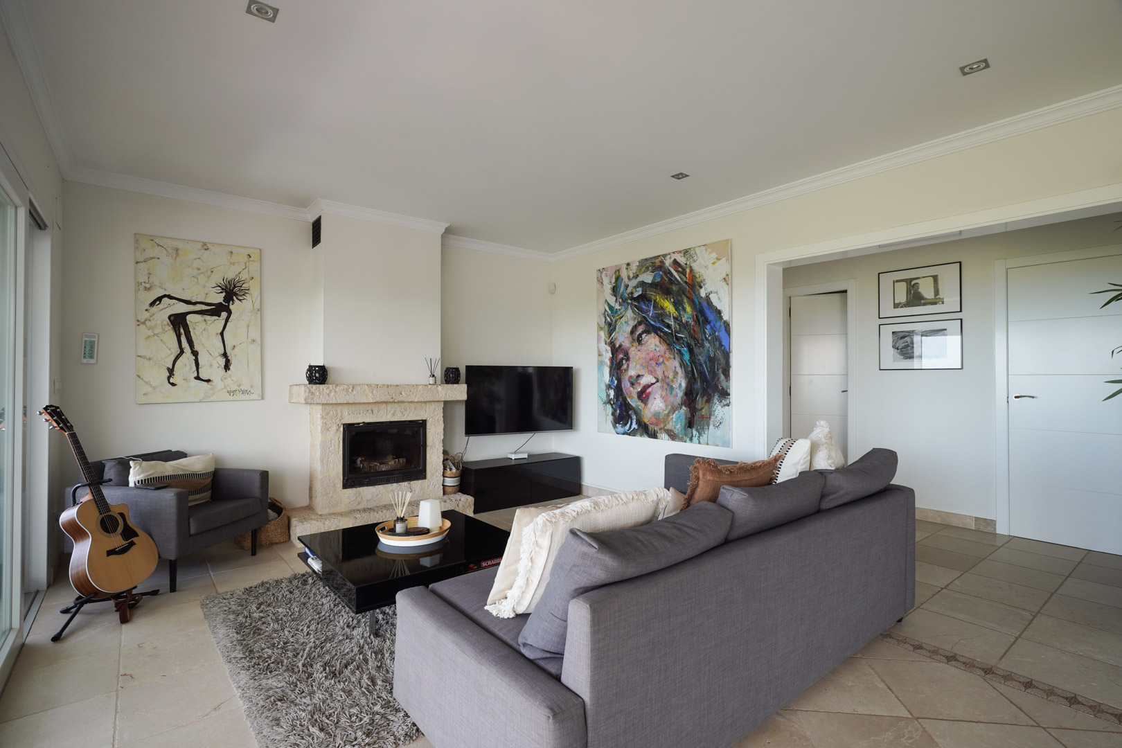 Espace de vie avec cheminée en pierre, canapés gris et sol carrelé avec frise décorative. Des œuvres d'art moderne et un éclairage encastré créent une ambiance raffinée.