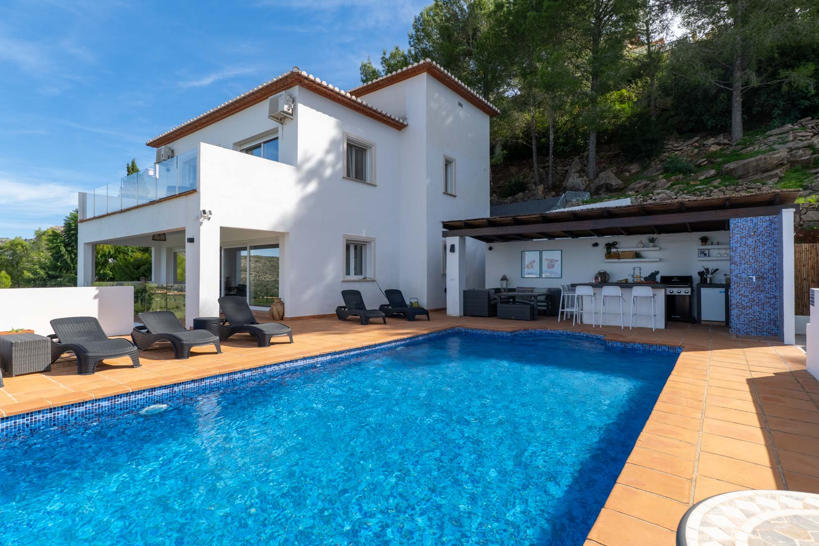 Espace extérieur avec piscine, terrasse en terre cuite et cuisine d'été couverte. La villa blanche de style méditerranéen est entourée d'une pinède et de parois rocheuses.
