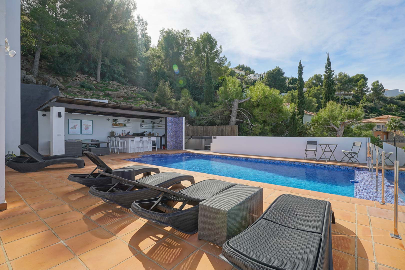 Espace piscine avec terrasse en terre cuite, bains de soleil en rotin et cuisine d'été sous pergola, offrant un cadre privé entouré de pins méditerranéens.