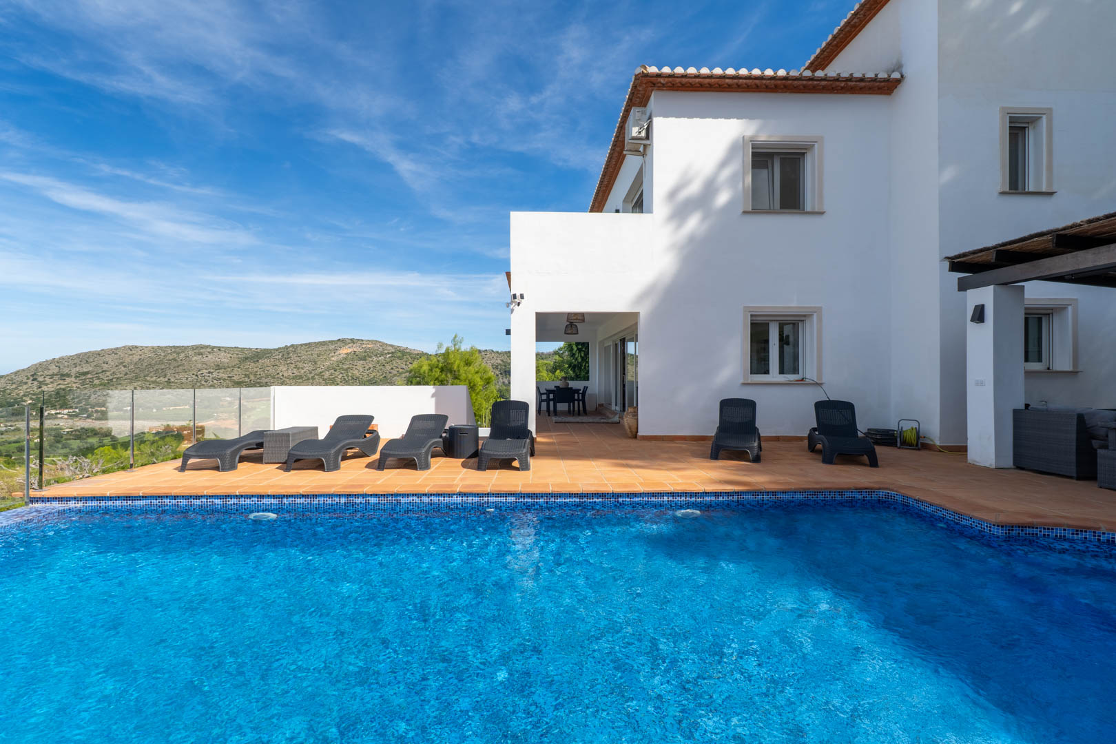 Piscine privée avec mosaïque bleue, entourée d'une terrasse en terre cuite avec chaises longues et vue dégagée sur les montagnes environnantes.