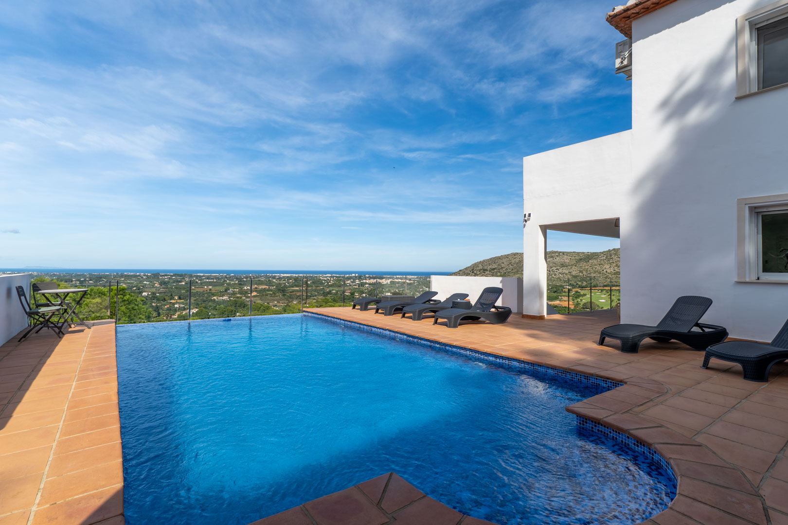 Piscine privée avec terrasse en terre cuite, bains de soleil noirs et garde-corps en verre offrant une vue panoramique sur la mer Méditerranée et le golf.
