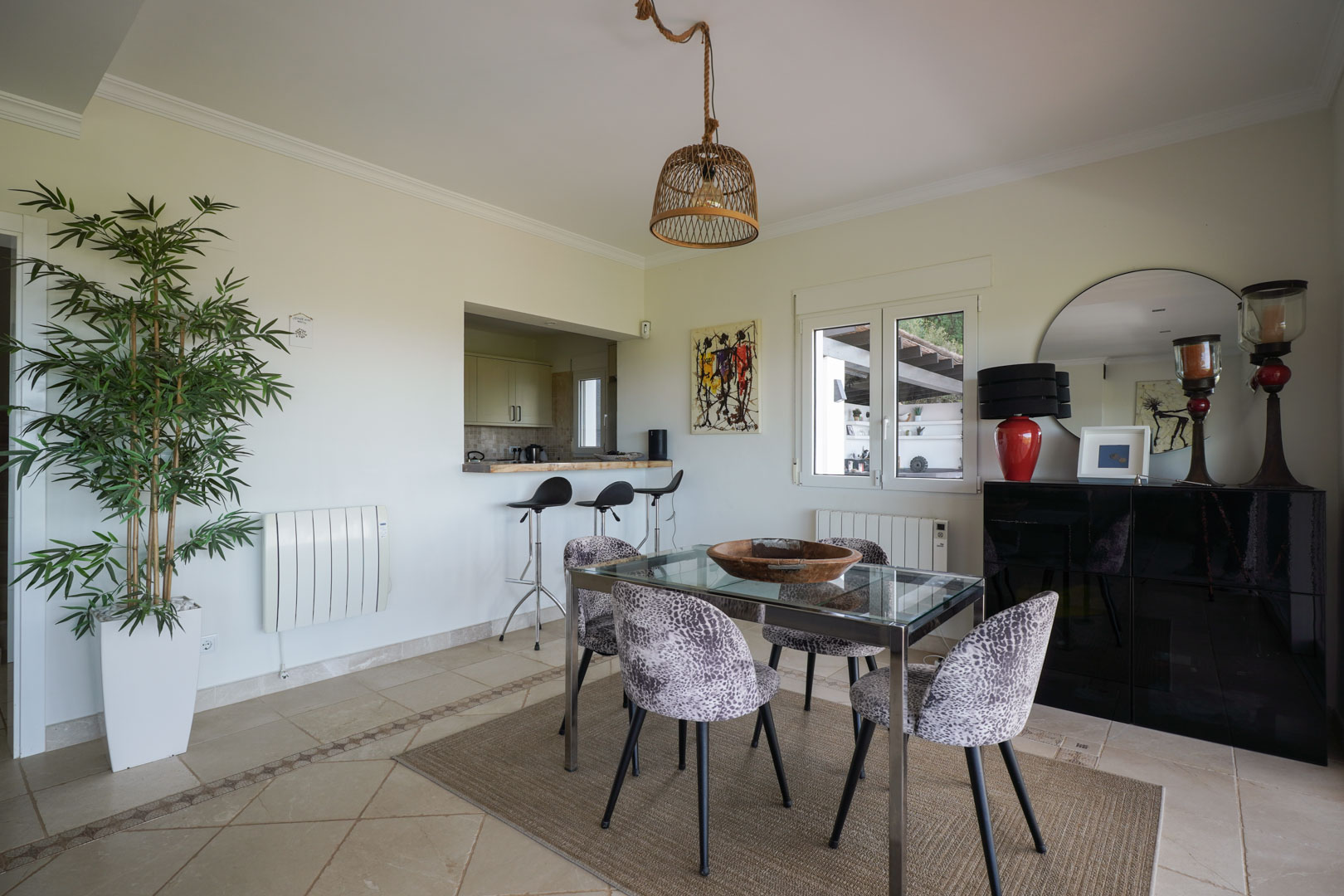 Salle à manger lumineuse avec table en verre, chaises à motif et suspension en osier, ouverte sur la cuisine par un comptoir de bar fonctionnel.