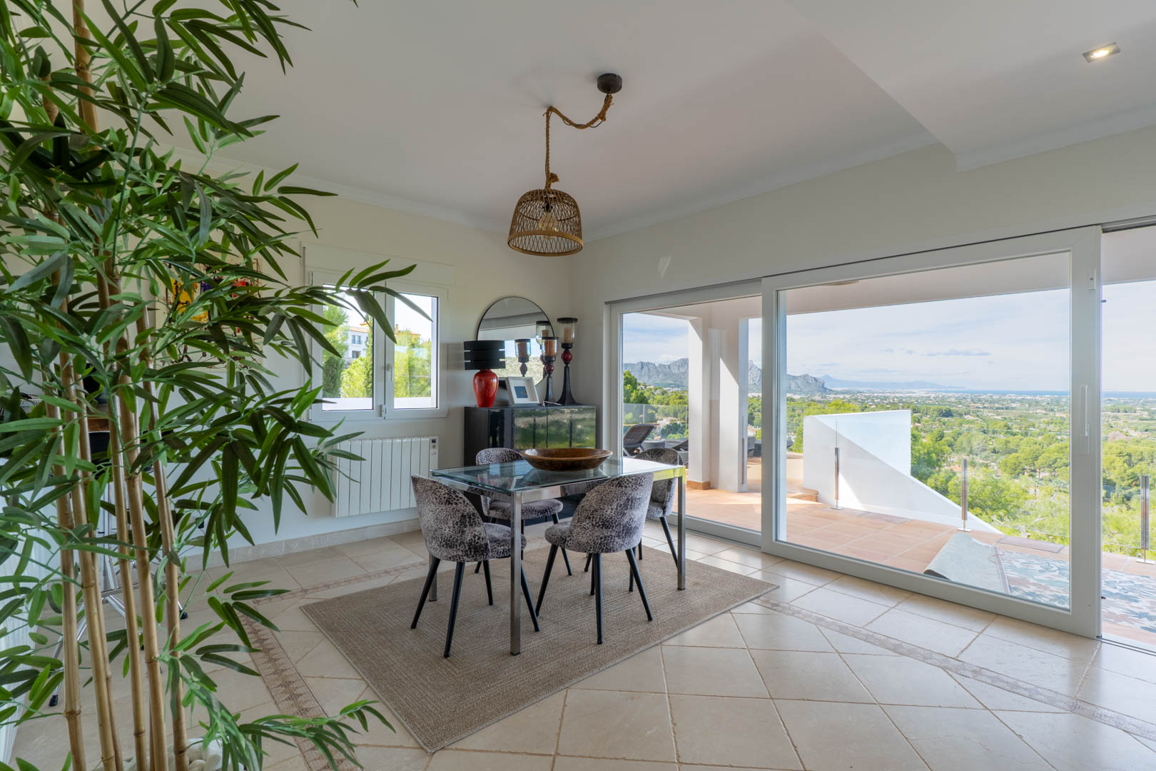Salle à manger lumineuse avec table en verre et grandes baies vitrées ouvrant sur une terrasse avec vue panoramique sur la mer et les montagnes.