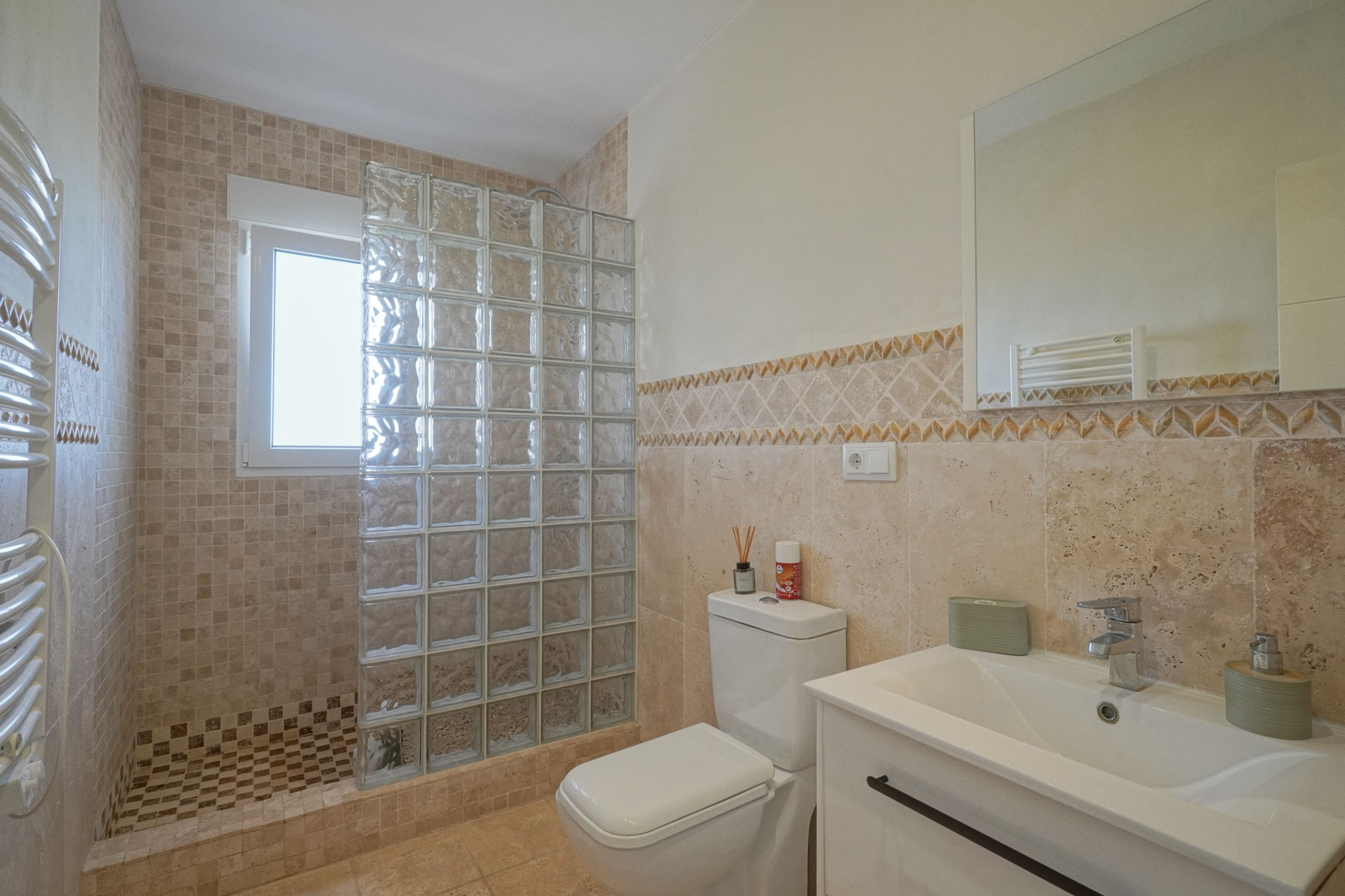 Salle de bains avec douche à l'italienne, paroi en briques de verre, carrelage aux tons naturels, meuble vasque blanc et radiateur sèche-serviettes.