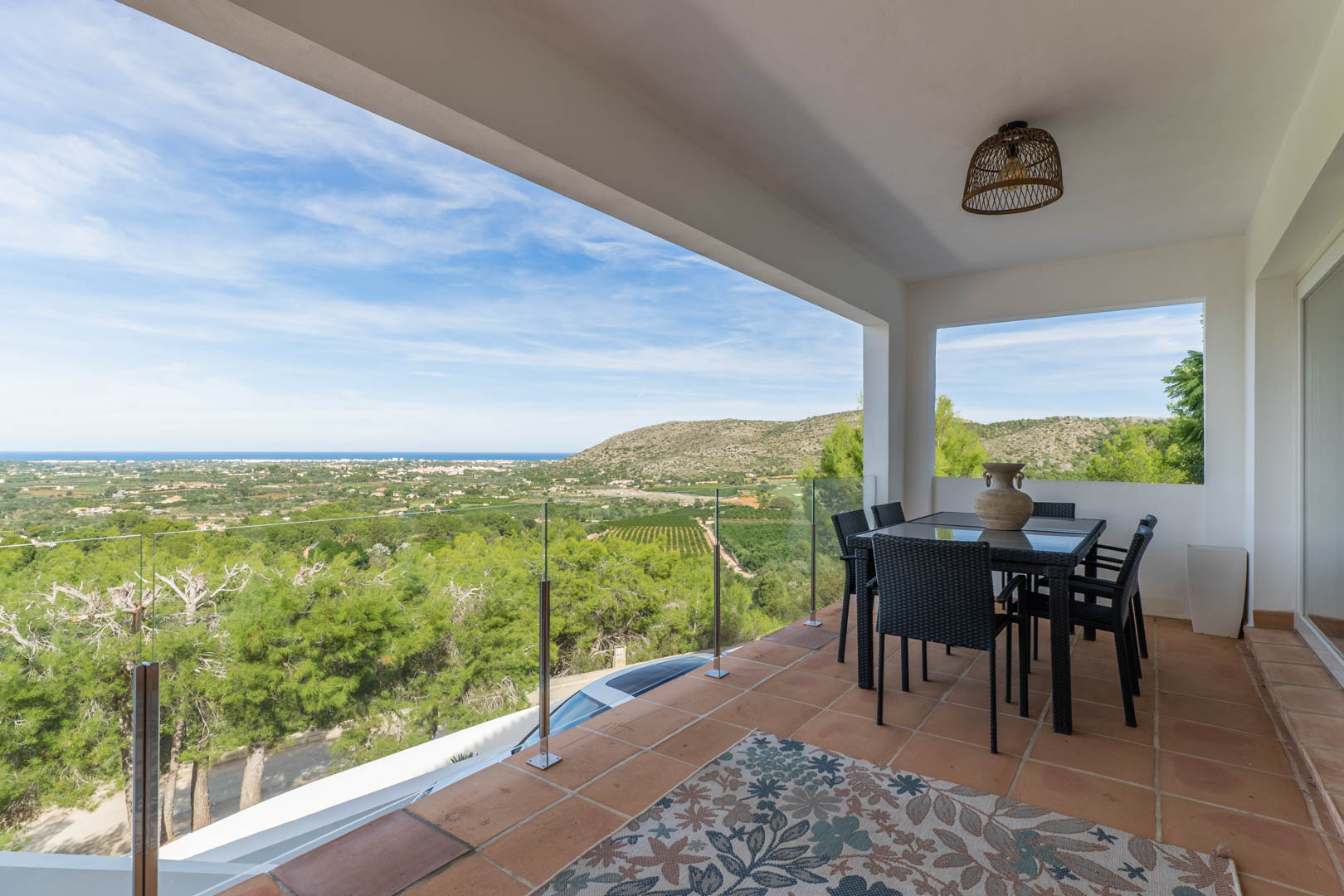 Terrasse couverte avec sol en terre cuite, mobilier de repas et garde-corps en verre offrant une vue panoramique sur la mer Méditerranée et la vallée.