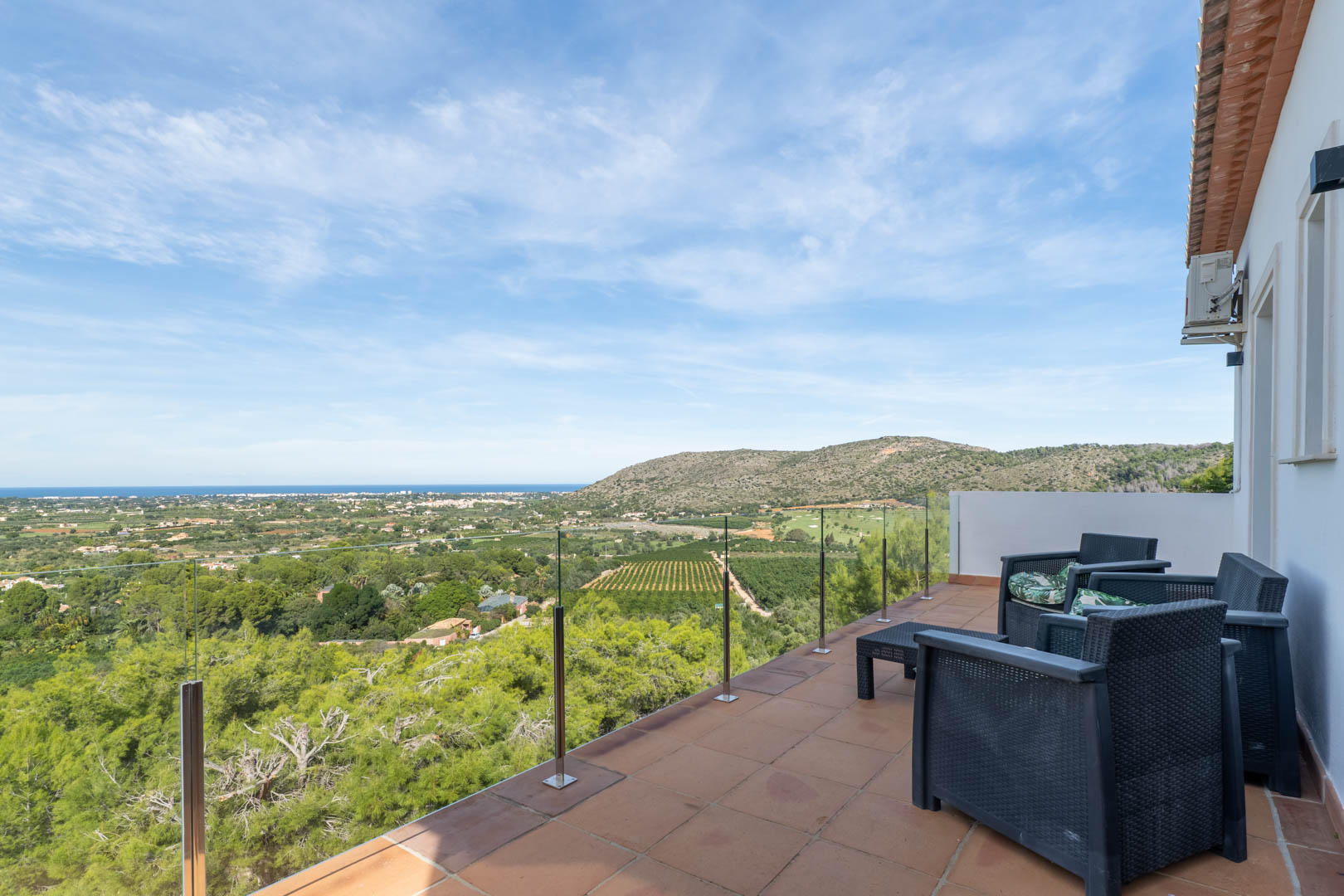 Terrasse supérieure avec garde-corps en verre, mobilier en résine tressée et vue panoramique sur la mer Méditerranée, le golf et les paysages naturels environnants.