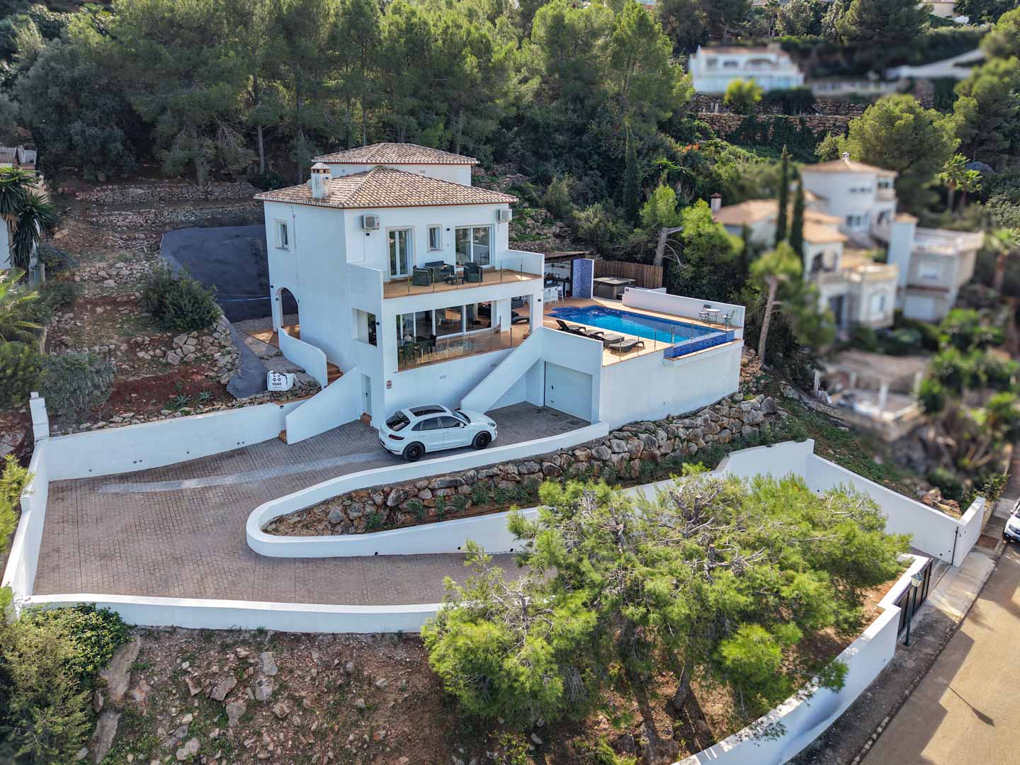 Vue aérienne de la villa avec façade blanche, piscine privée, terrasses vitrées et allée pavée, le tout niché dans l'environnement boisé de La Sella.