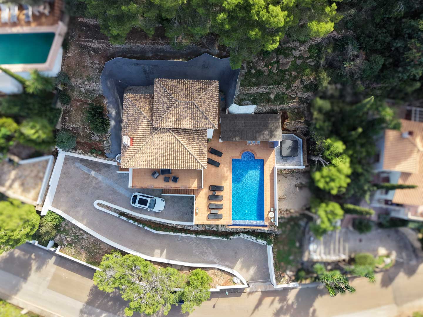 Vue aérienne de la villa montrant le toit en tuiles, la piscine rectangulaire, la terrasse avec bains de soleil, le jacuzzi extérieur et l'allée de stationnement privée.
