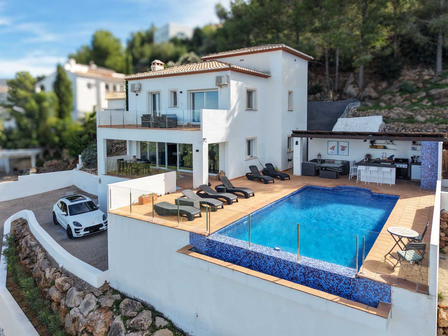 Vue extérieure de la villa avec piscine en mosaïque bleue, solarium, cuisine d'été sous pergola et façade blanche méditerranéenne sur un terrain en pente paysagé.