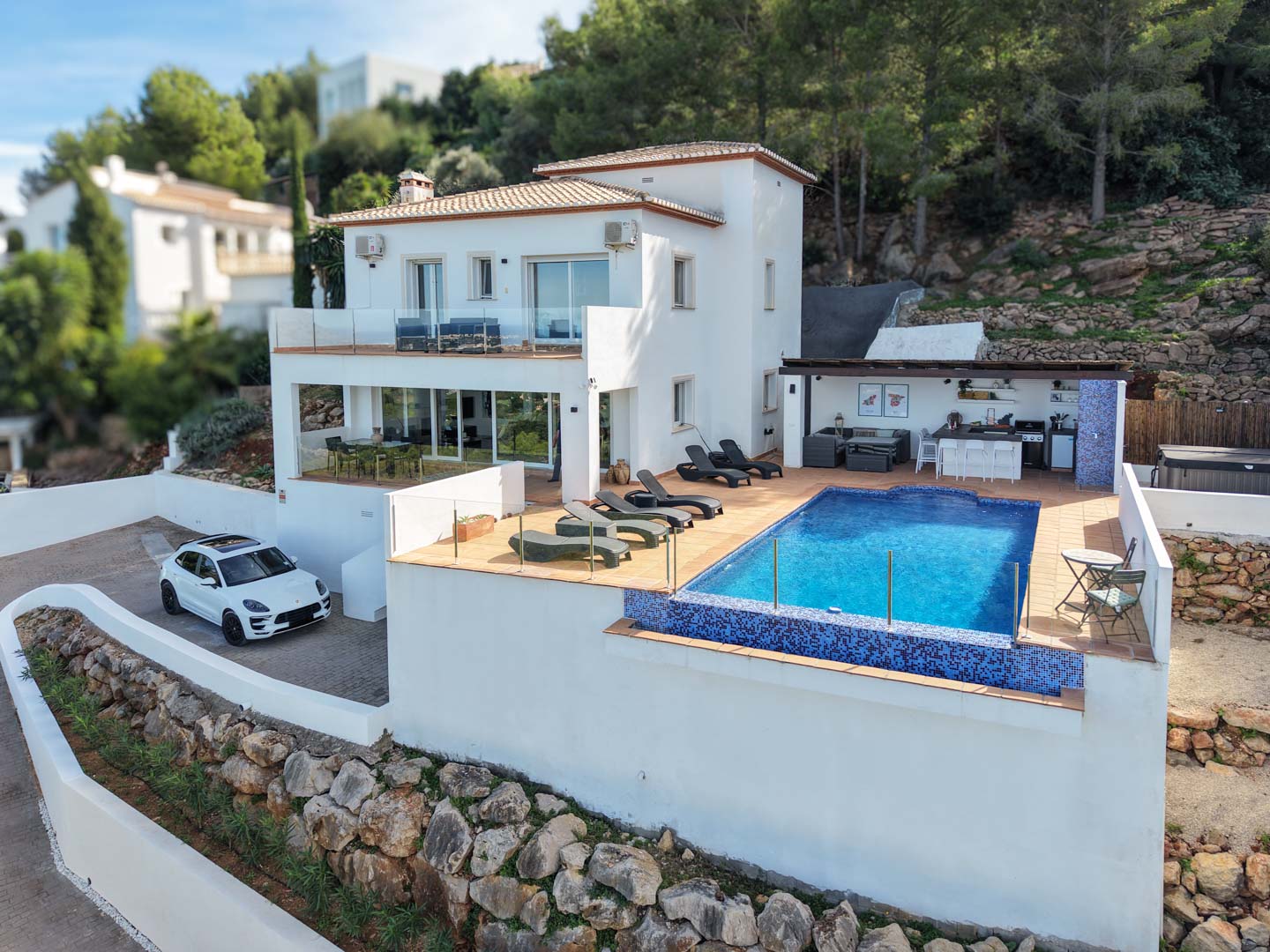 Vue extérieure de la villa avec piscine en mosaïque bleue, terrasse ensoleillée et cuisine d'été équipée, le tout niché dans un environnement naturel préservé.