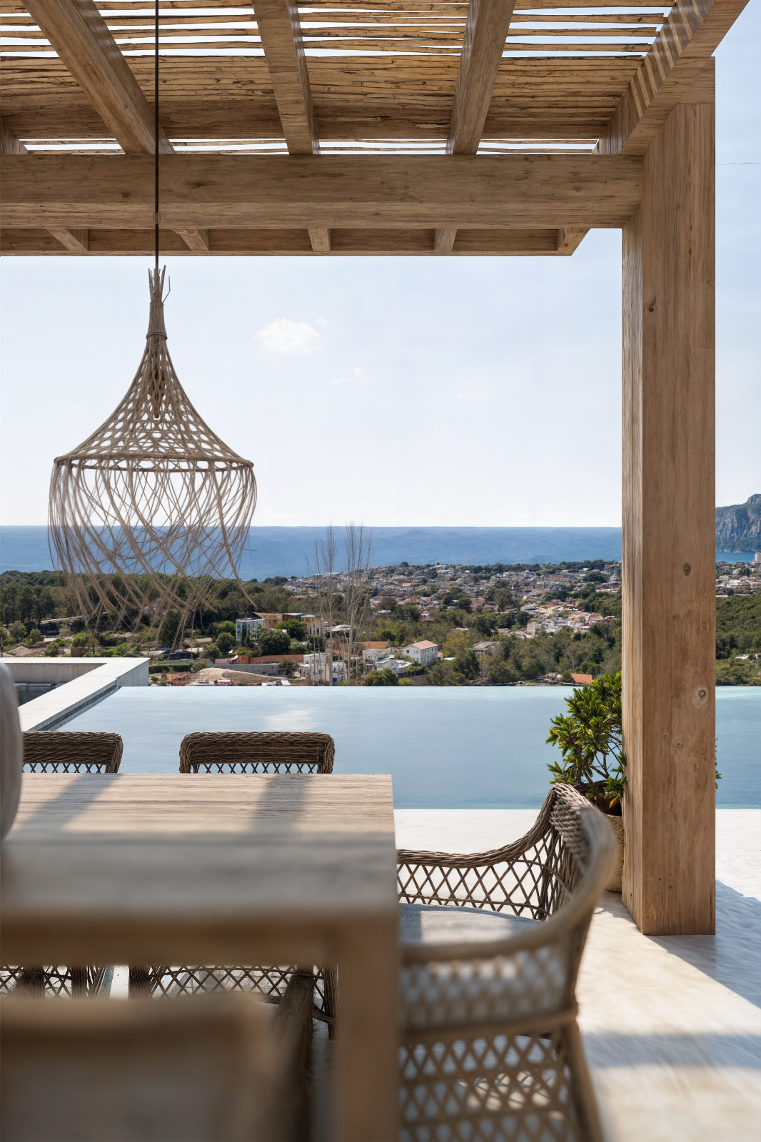 Terrasse extérieure avec pergola en bois, salle à manger en osier et luminaire tressé offrant une vue panoramique sur la piscine à débordement et la mer.
