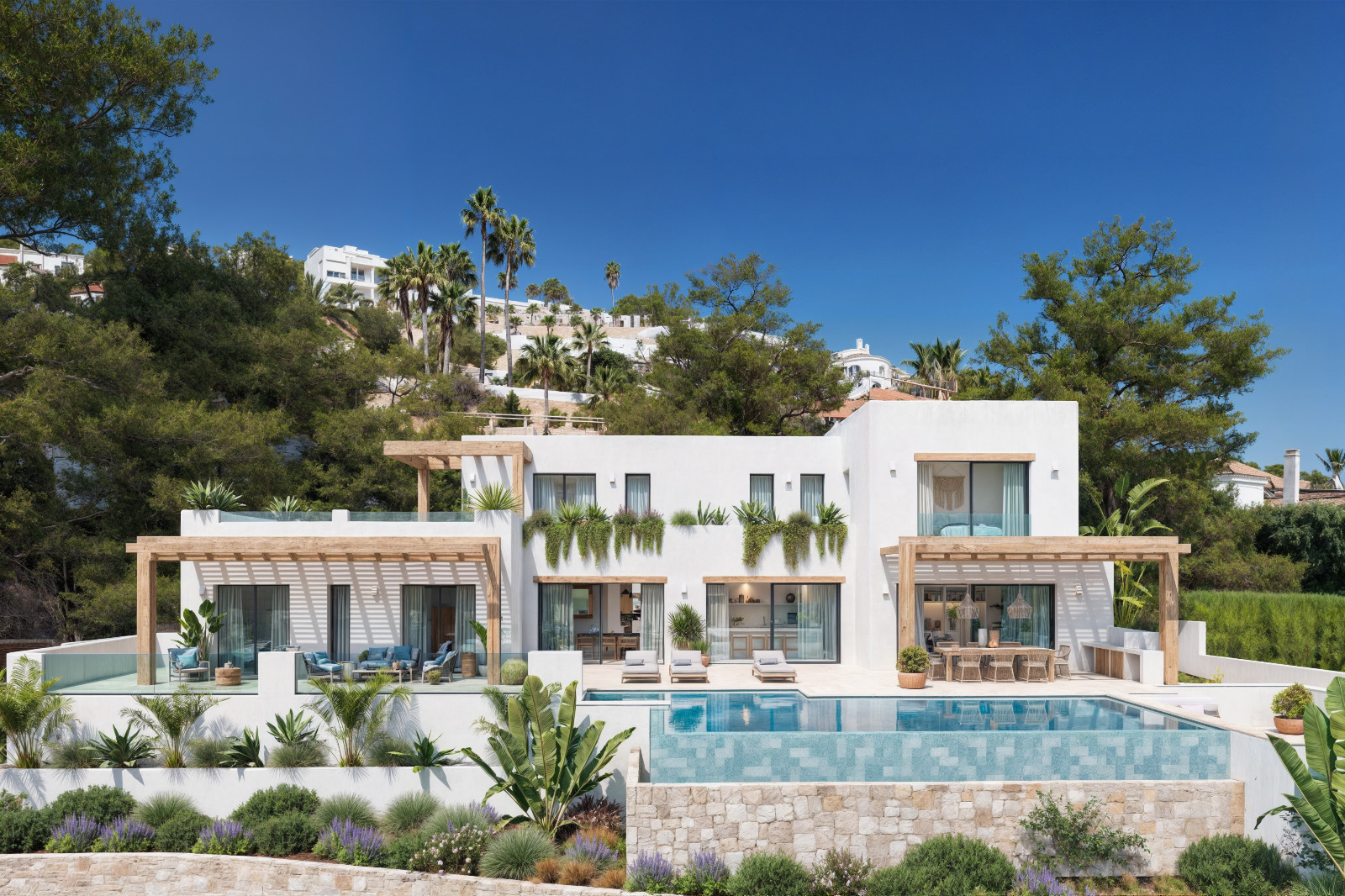 Vue extérieure de la villa avec façades blanches, pergolas en bois, piscine à débordement et jardin méditerranéen avec murs en pierre naturelle.