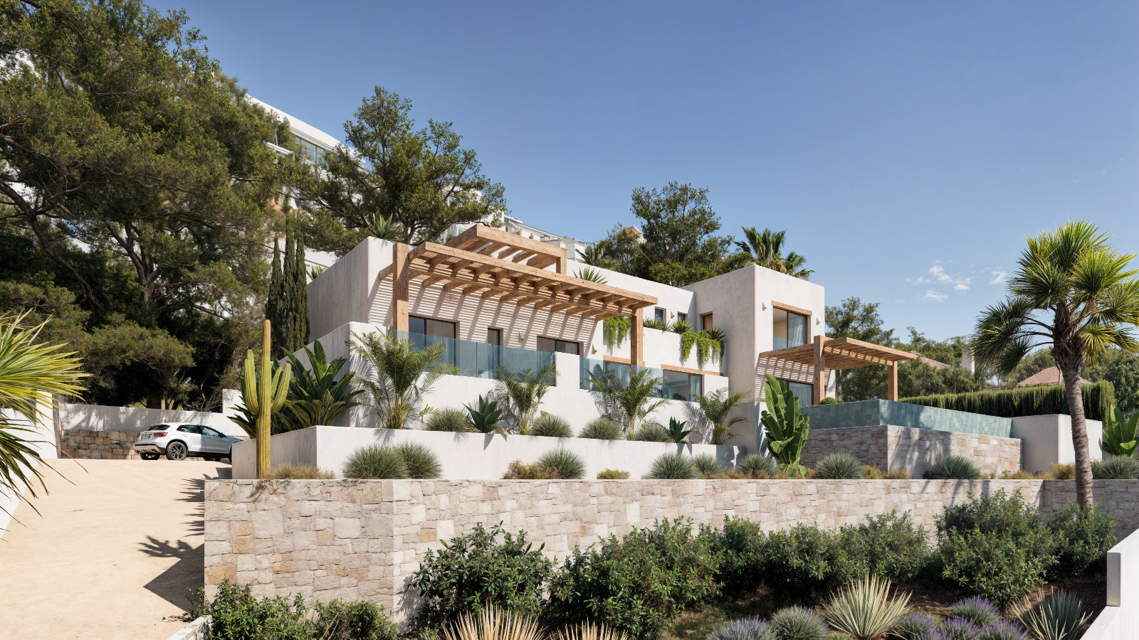 Vue extérieure de la villa de style ibizane avec façades blanches, pergolas en bois et murs en pierre naturelle intégrés dans un jardin méditerranéen avec palmiers.