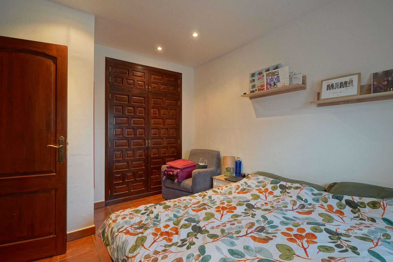 Chambre avec placard intégré en bois sculpté, sol en terre cuite et étagères murales. L'éclairage par spots encastrés souligne le style méditerranéen classique.