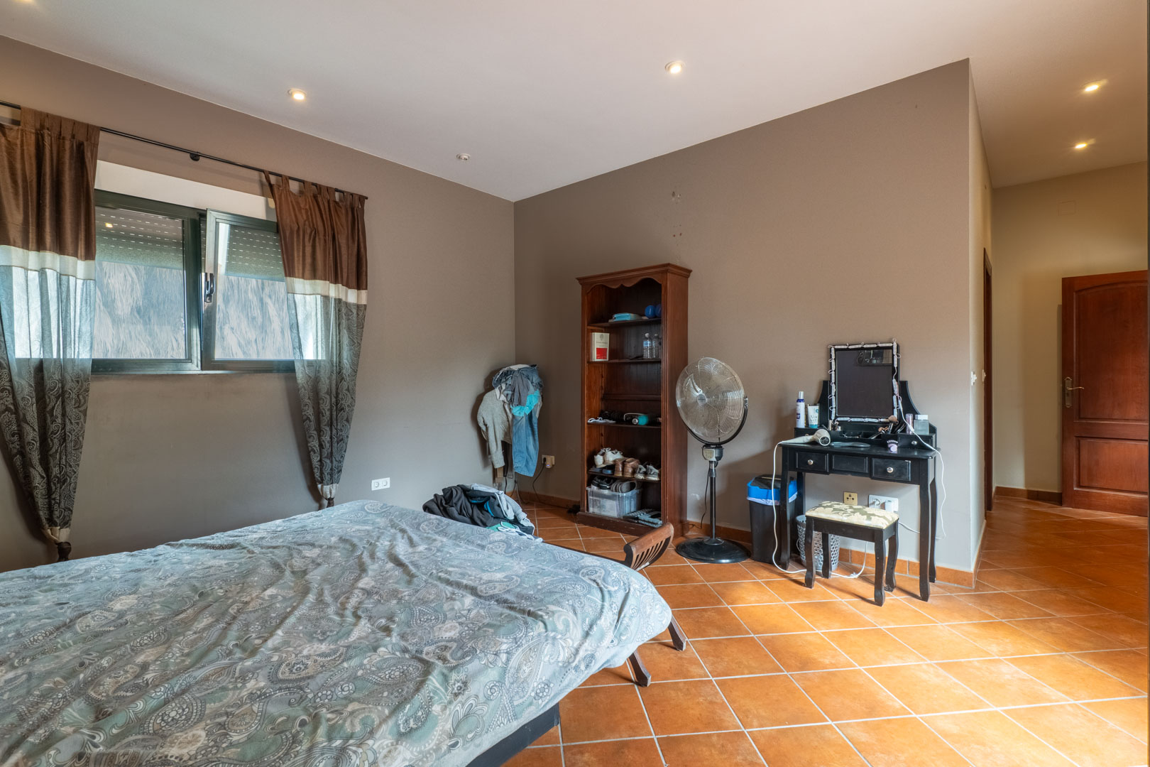Chambre avec sol en terre cuite, éclairage intégré au plafond et murs taupe, comprenant une fenêtre avec volets et une porte en bois sombre.