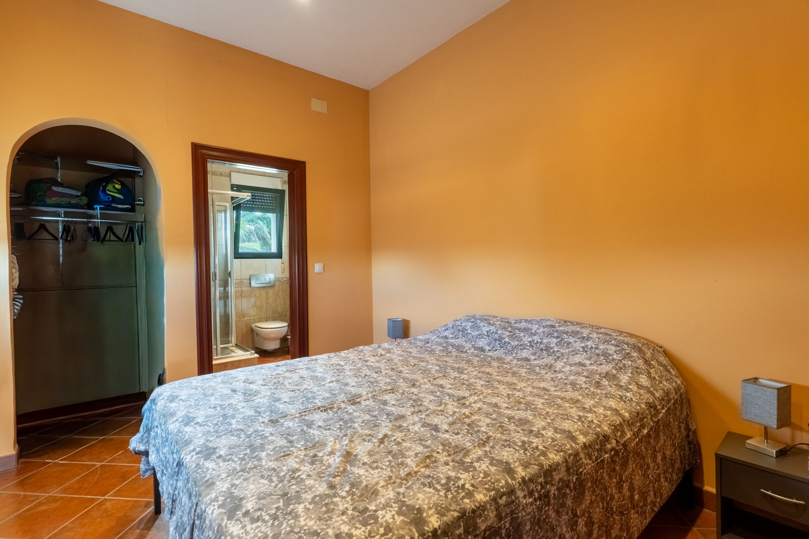 Chambre avec sol en terre cuite et murs ocre, comprenant un dressing voûté et une salle de bain attenante avec cabine de douche.