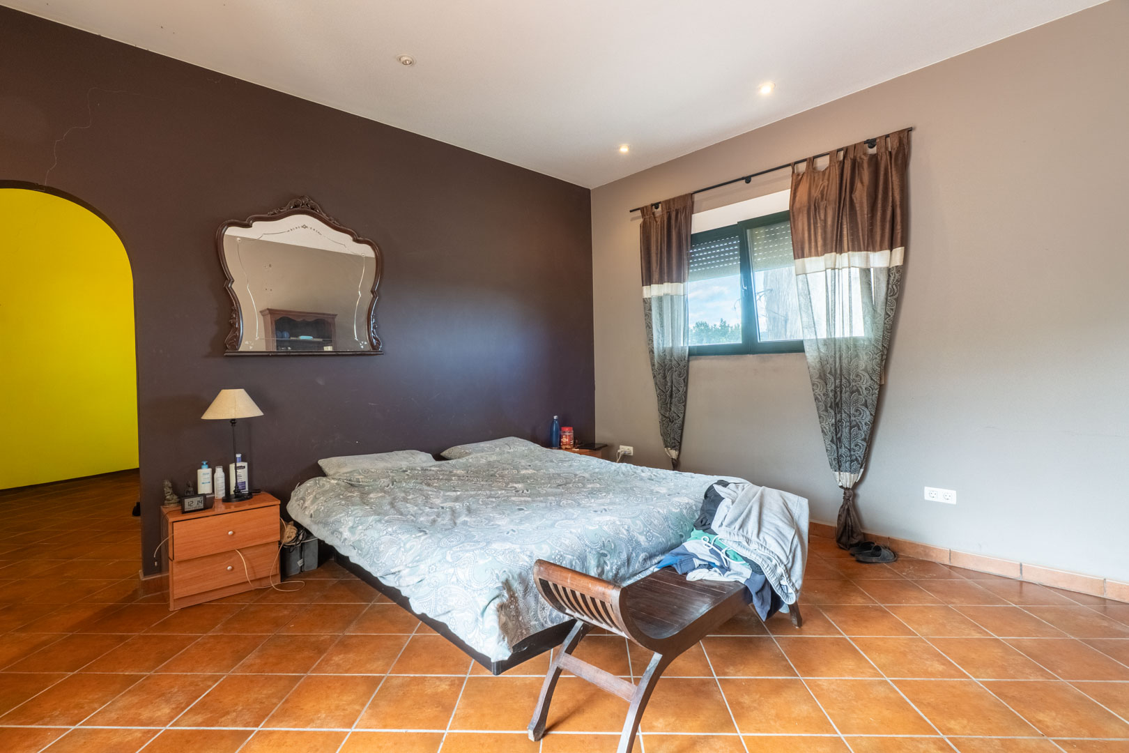 Chambre avec sol en terre cuite, mur d'accent marron foncé et miroir ancien. Une ouverture en arche mène à une pièce adjacente sous un éclairage encastré.