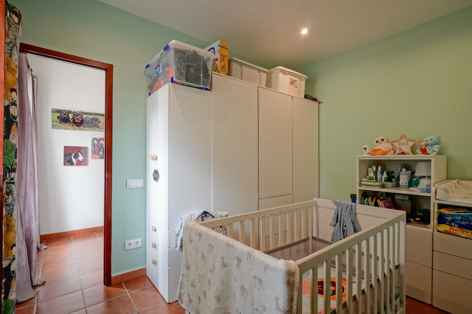 Chambre d'enfant avec berceau en bois blanc, armoires de rangement et murs vert pastel. Le sol est en terre cuite traditionnelle, complété par un éclairage encastré.