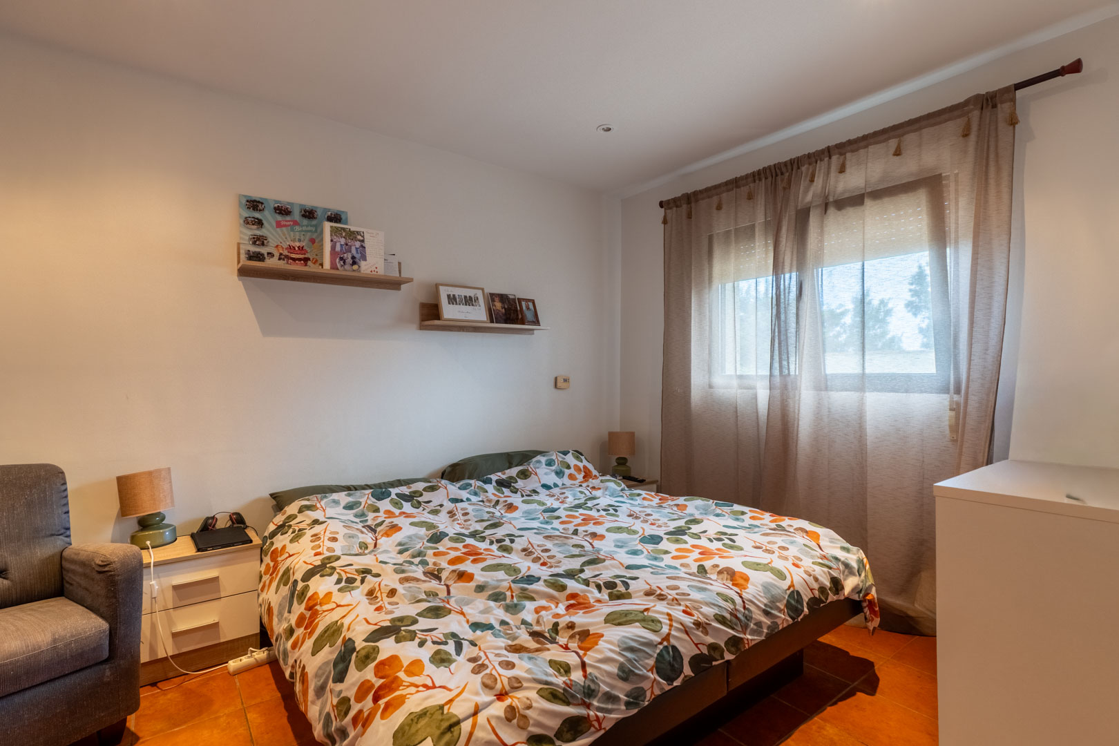 Chambre lumineuse avec sol en terre cuite, murs blancs et étagères en bois. Une fenêtre avec rideaux translucides et volet roulant assure une belle luminosité naturelle.