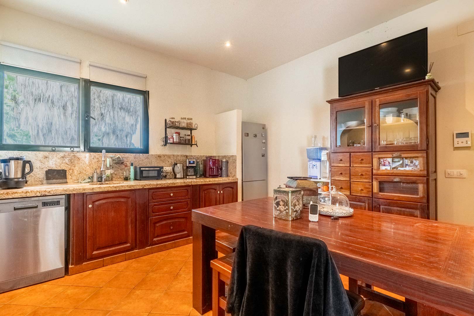 Cuisine rustique avec plans de travail en granit, meubles en bois sombre et table à manger centrale. Équipée d'appareils en inox et éclairée par une large fenêtre donnant sur l'extérieur.