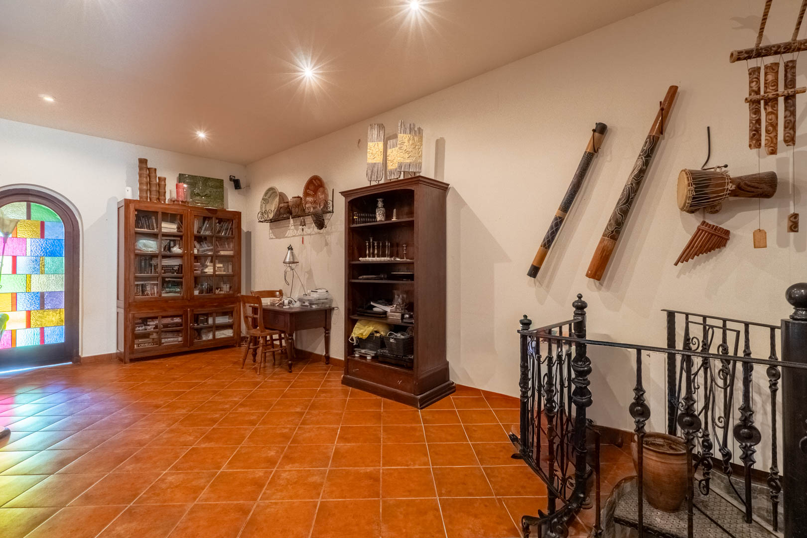 Espace bureau avec sol en terre cuite, bibliothèques en bois, instruments ethniques et accès à l'escalier en colimaçon avec rampe en fer forgé.