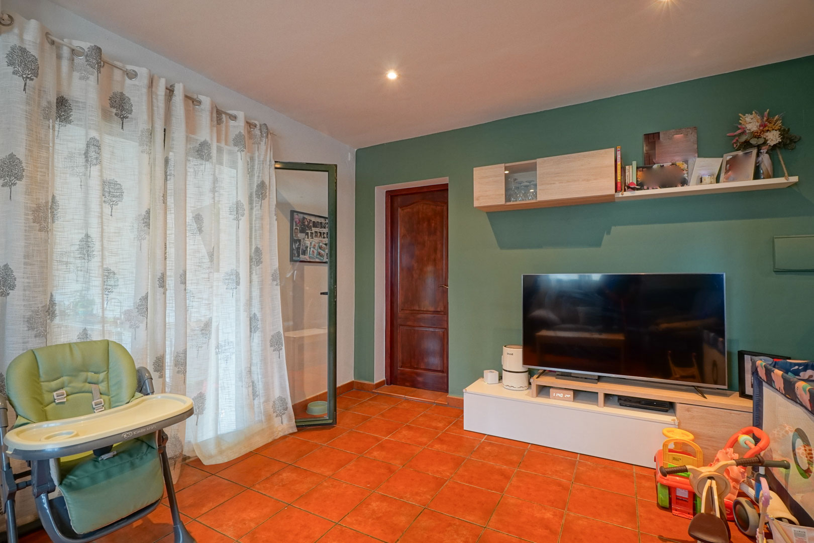Espace de vie avec mur d'accent vert sauge, sol en terre cuite et meuble TV moderne. Une porte en bois massif et des spots encastrés complètent cette pièce lumineuse.