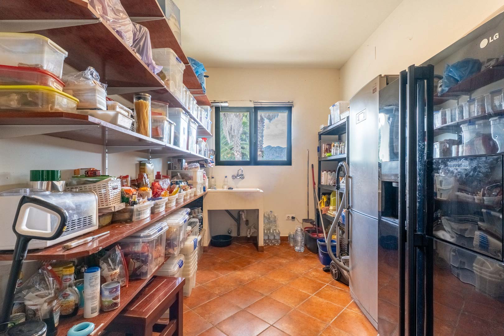 Garde-manger fonctionnel avec de grandes étagères en bois, un évier sous la fenêtre offrant une vue dégagée et des réfrigérateurs sur un sol en terre cuite.