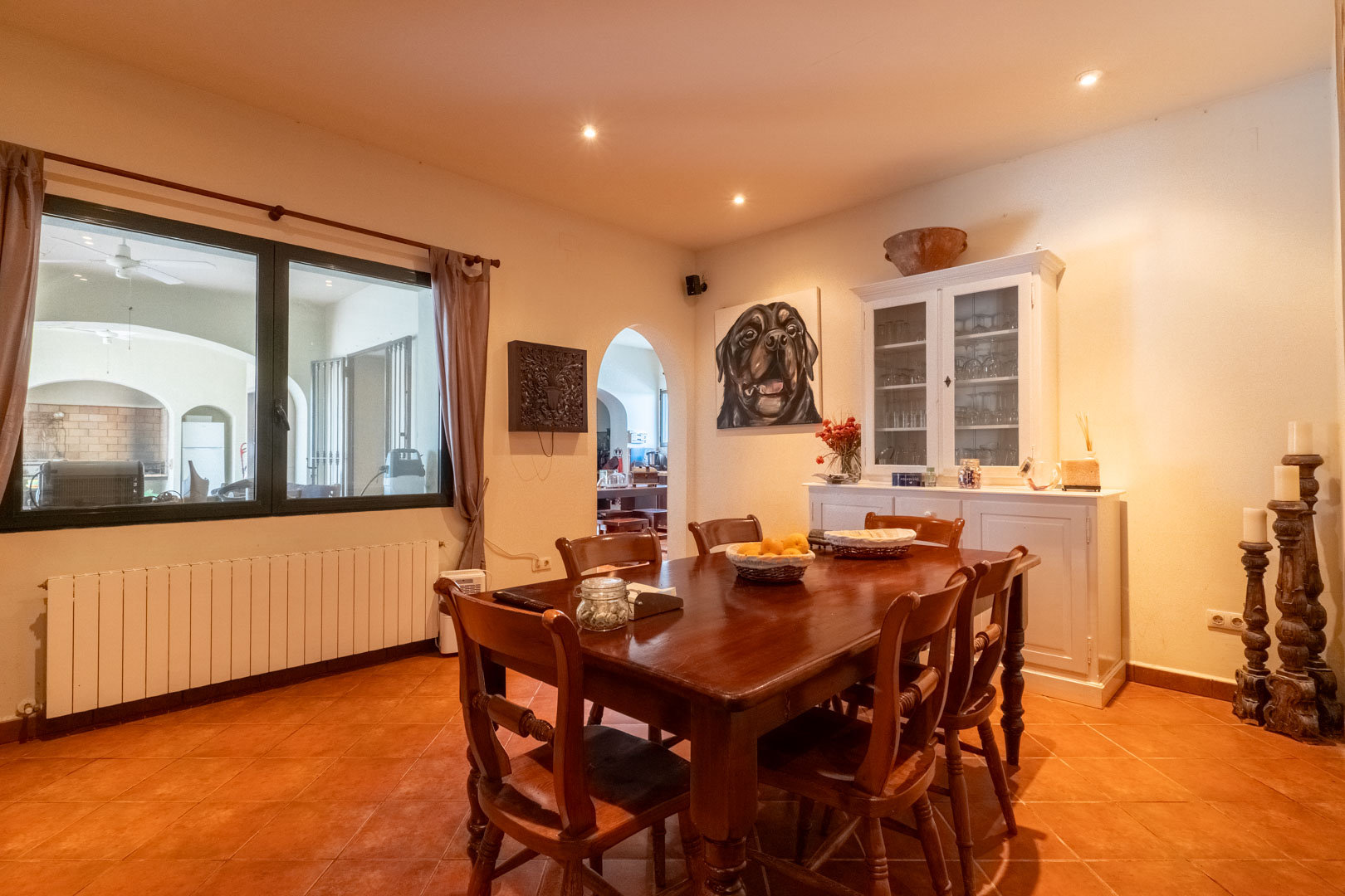 Salle à manger avec sol en terre cuite, table en bois massif et buffet blanc. Une grande fenêtre offre une vue sur le patio couvert, sous un éclairage encastré.