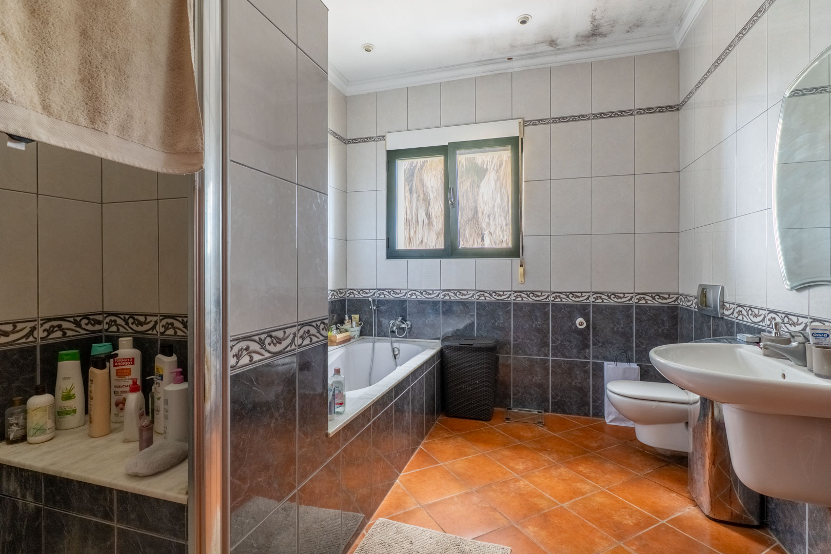 Salle de bain avec carrelage bicolore, baignoire encastrée et sol en terre cuite. Équipée d'un WC suspendu, d'un lavabo sur colonne et d'un éclairage encastré.