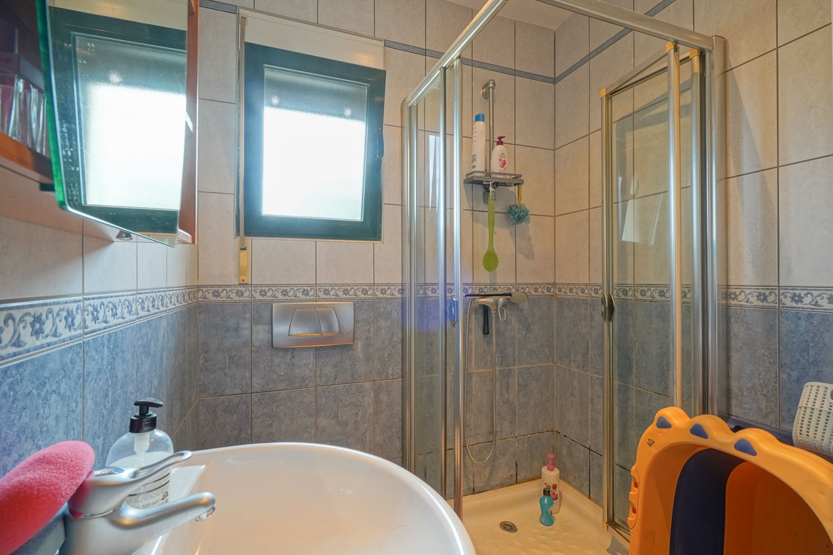 Salle d'eau fonctionnelle avec cabine de douche en verre, lavabo en céramique blanche et carrelage mural bleu orné d'une frise décorative et d'une fenêtre.
