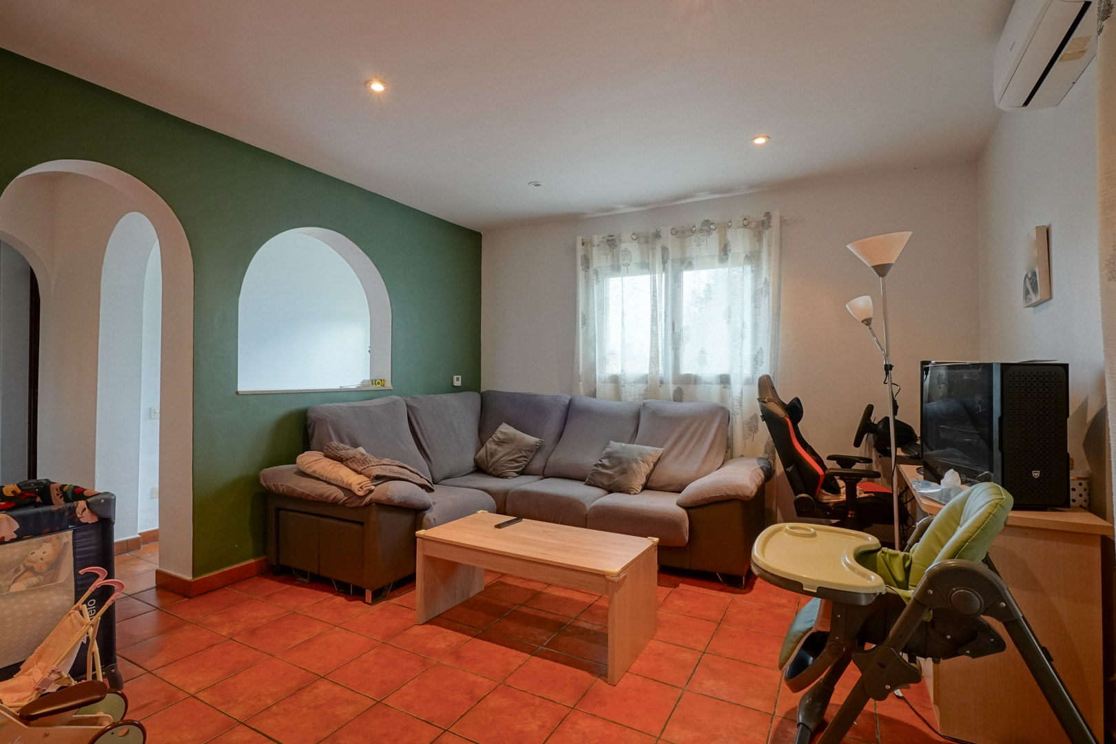 Salon avec sol en terre cuite, mur d'accent vert et ouvertures en arche. Comprend un canapé d'angle, un coin bureau et la climatisation réversible.