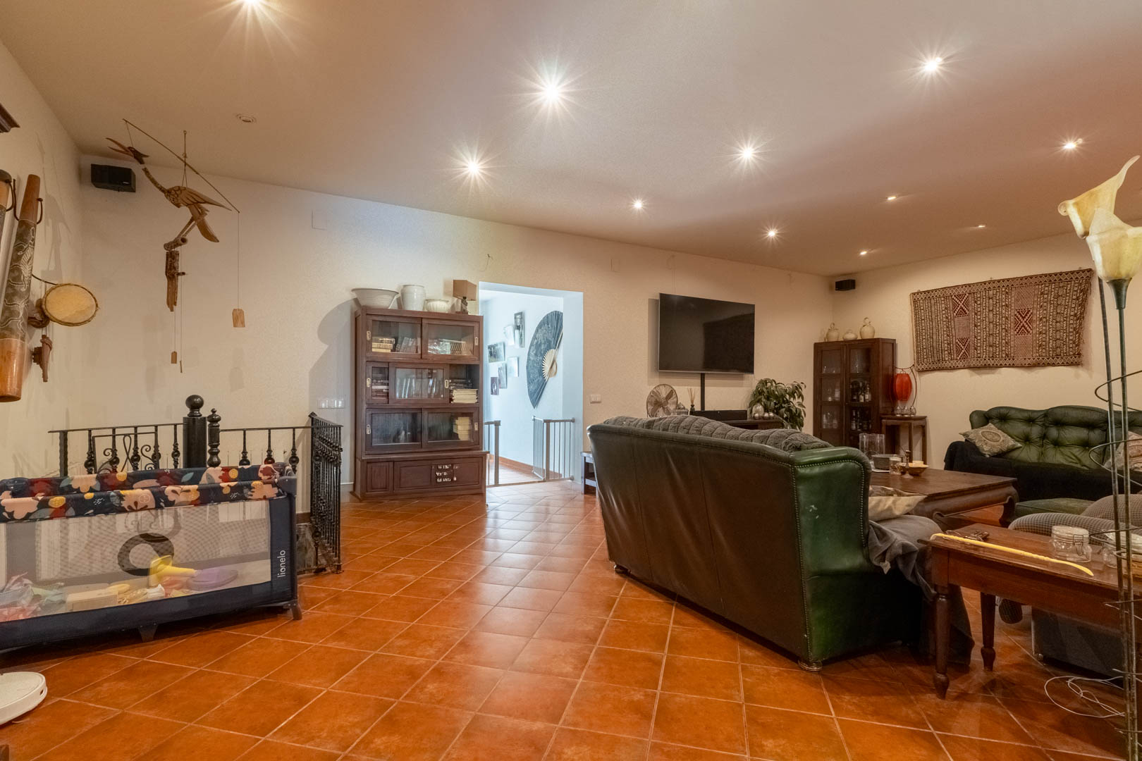 Salon spacieux avec sol en terre cuite, canapés en cuir vert et mobilier en bois classique. L'espace dispose d'un éclairage encastré et d'un escalier en fer forgé.