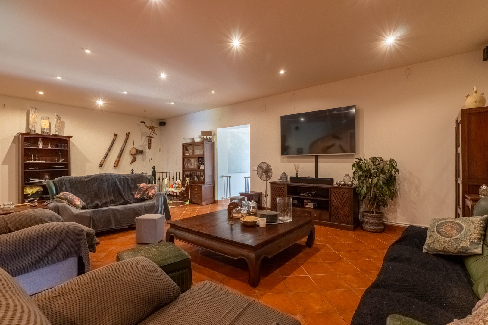 Salon spacieux avec sol en terre cuite, éclairage encastré au plafond et mobilier en bois massif. L'espace comprend un coin multimédia et un accès à l'escalier en fer forgé.
