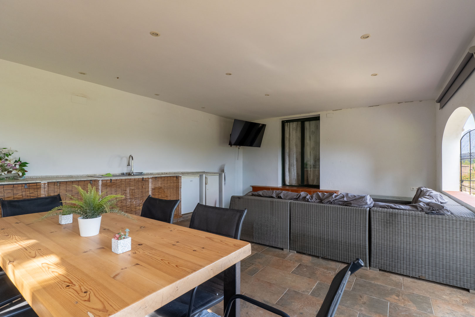 Terrasse couverte avec salon en rotin gris, table à manger en bois massif et kitchenette extérieure. L'espace dispose de spots encastrés au plafond et d'un sol carrelé.