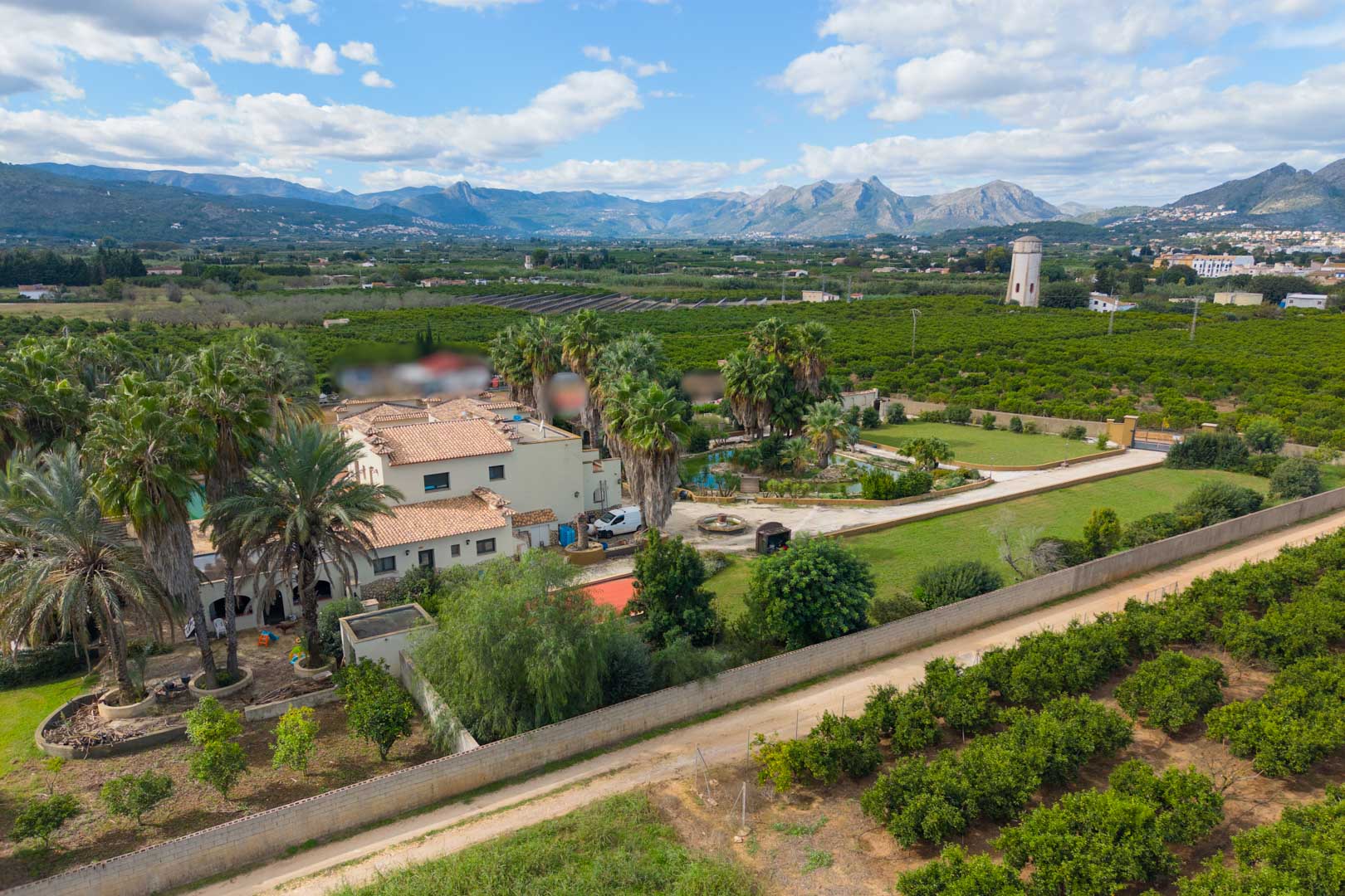 Vue aérienne de la propriété incluant la villa principale, les jardins de palmiers et l'étang, entourée de vergers avec les montagnes en arrière-plan.