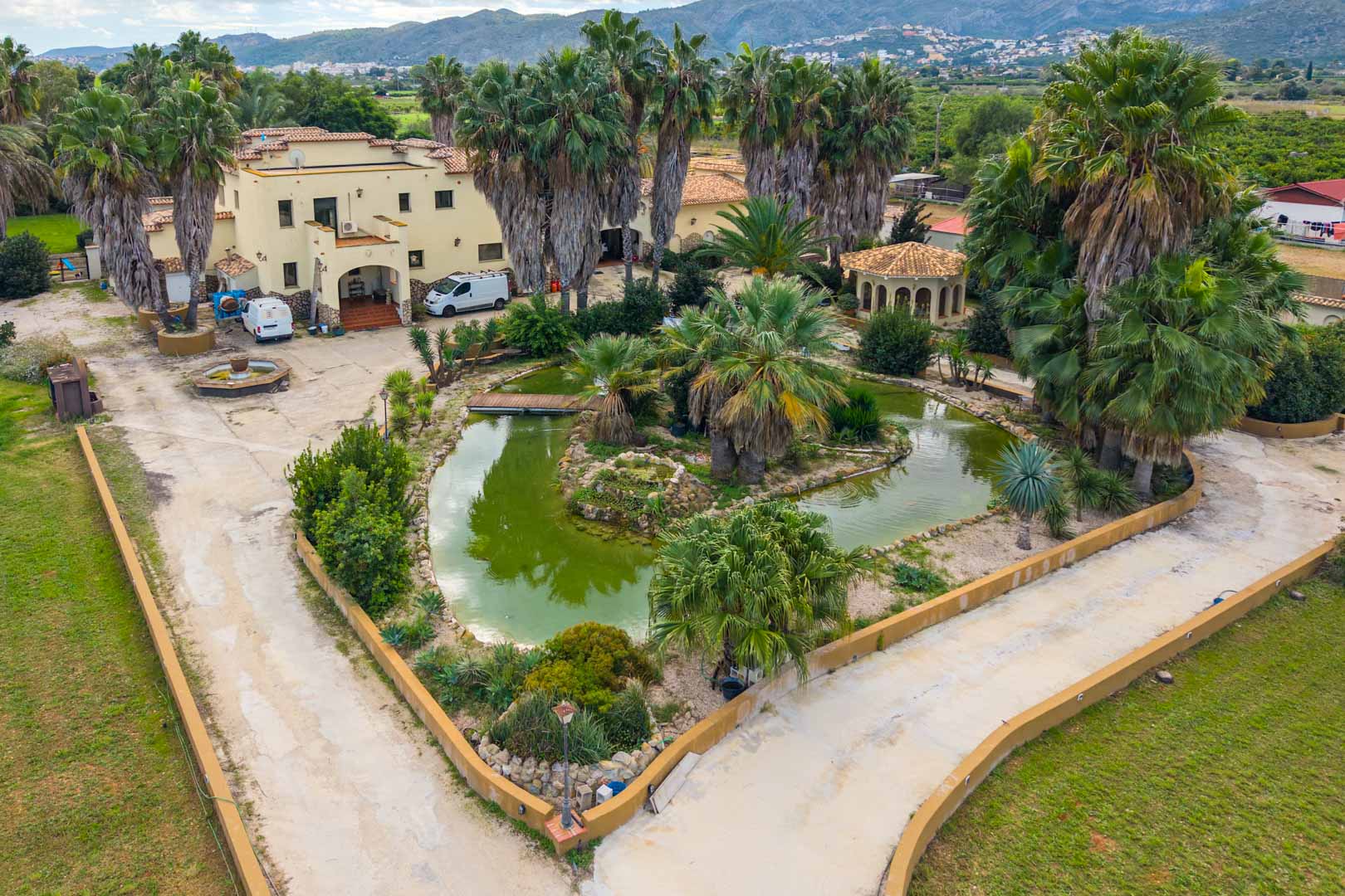 Vue aérienne de l'entrée principale avec un étang ornemental, un pont en bois et des palmiers matures devant la villa de style méditerranéen.