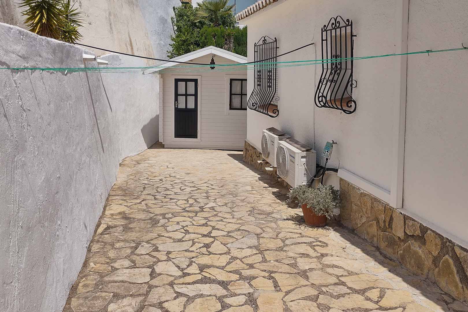 Allée latérale pavée de pierres naturelles menant à un abri de jardin en bois, bordée par la façade avec climatiseurs et grilles de fenêtres en fer forgé.