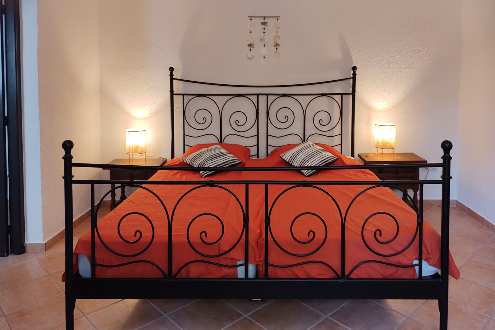Chambre avec cadre de lit en fer forgé noir, tables de chevet en bois et sol en grès cérame. L'éclairage tamisé souligne la texture des murs blancs.
