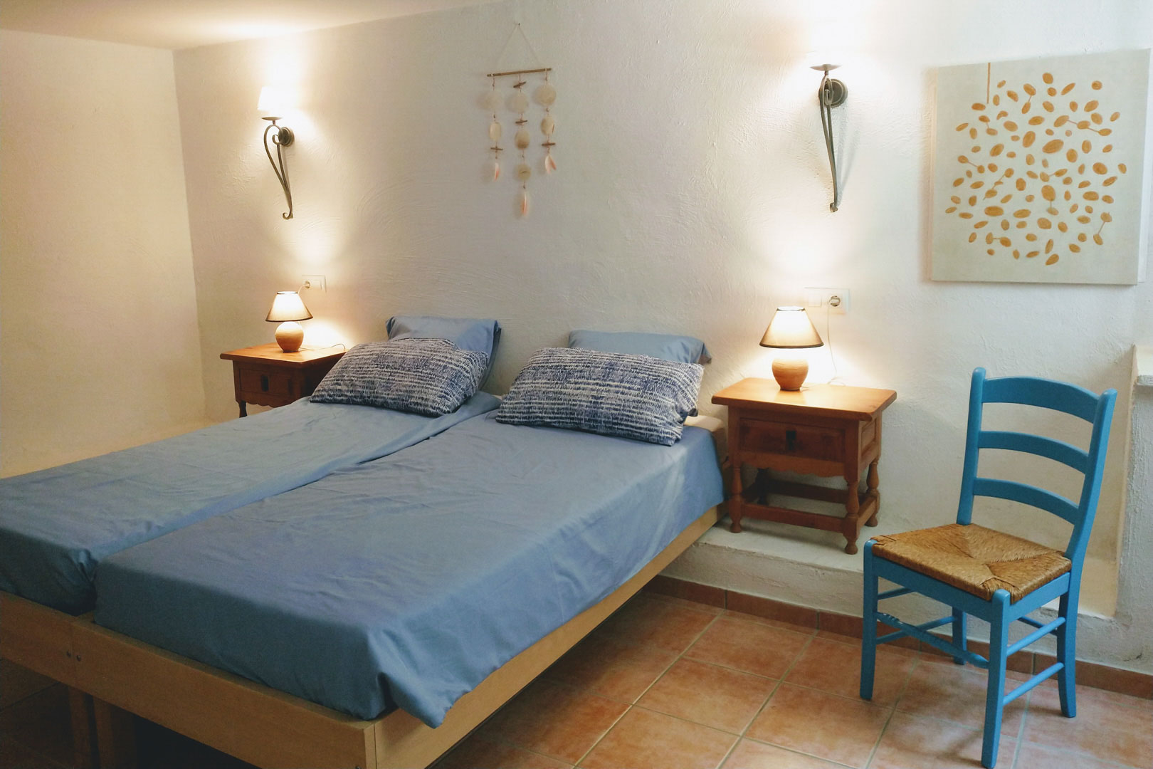 Chambre avec deux lits simples, sol en terre cuite et murs blancs texturés. Elle dispose de tables de chevet en bois, d'appliques murales et d'une chaise bleue.