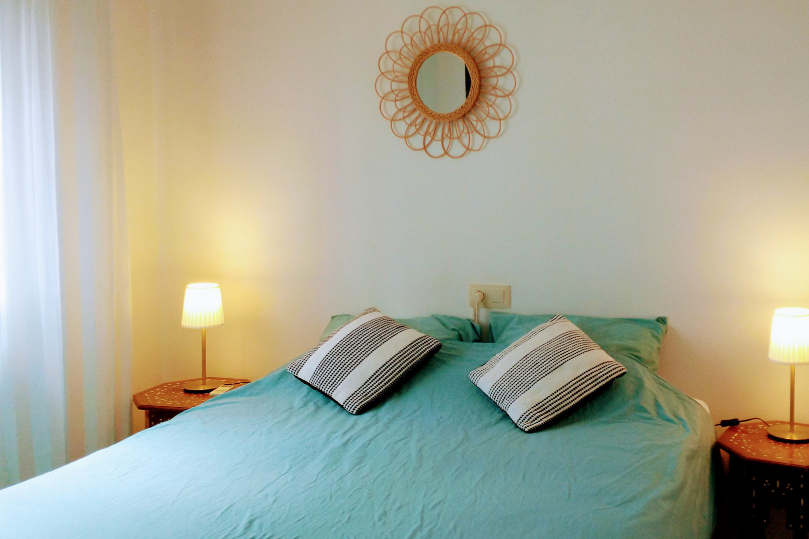 Chambre avec lit double, linge de lit turquoise, tables de chevet hexagonales en bois et miroir décoratif en rotin sur un mur blanc épuré.