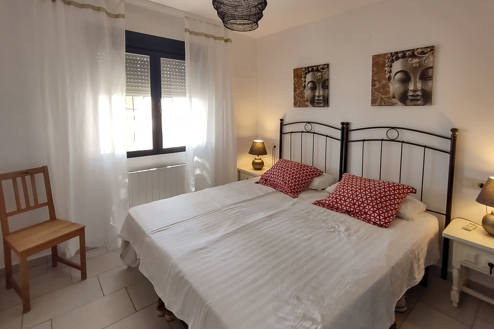 Chambre avec lits jumeaux, têtes de lit en fer forgé noir, tableaux de Bouddha et fenêtre avec volets roulants et rideaux blancs vaporeux.