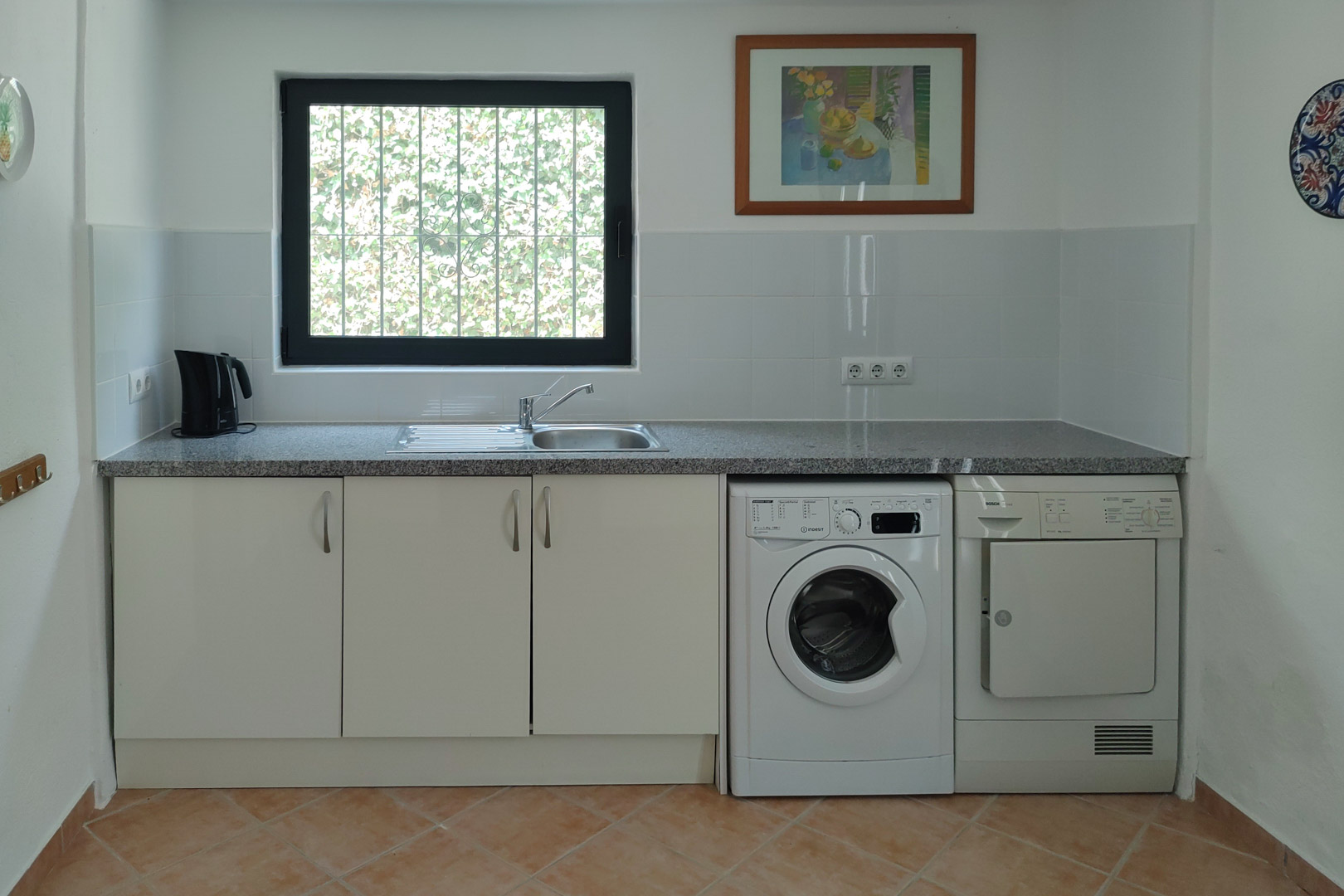 Espace buanderie pratique avec plan de travail en granit, évier en inox, lave-linge et sèche-linge encastrés sous des meubles blancs et sol en grès.