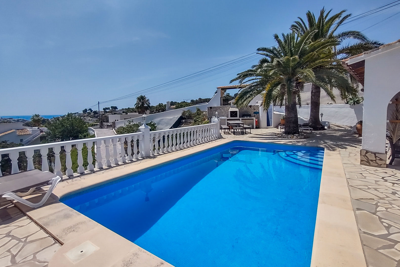 Espace piscine avec terrasse en pierre, balustrade blanche et vue mer. À l'arrière-plan, un coin barbecue avec palmiers et table de pique-nique.