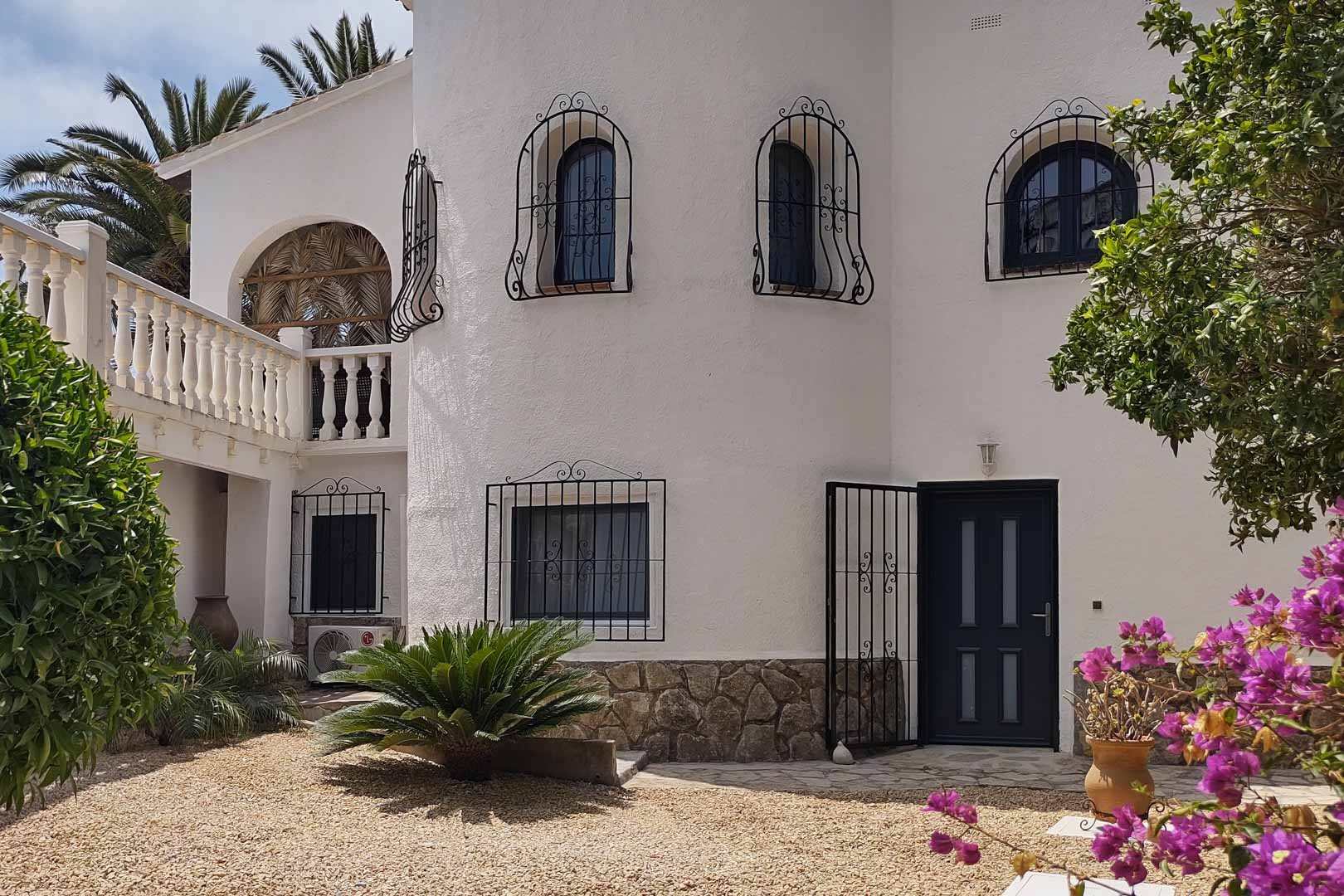 Façade méditerranéenne blanche avec soubassement en pierre, fenêtres ornées de grilles en fer forgé et entrée principale entourée d'un jardin de gravier et de bougainvilliers.
