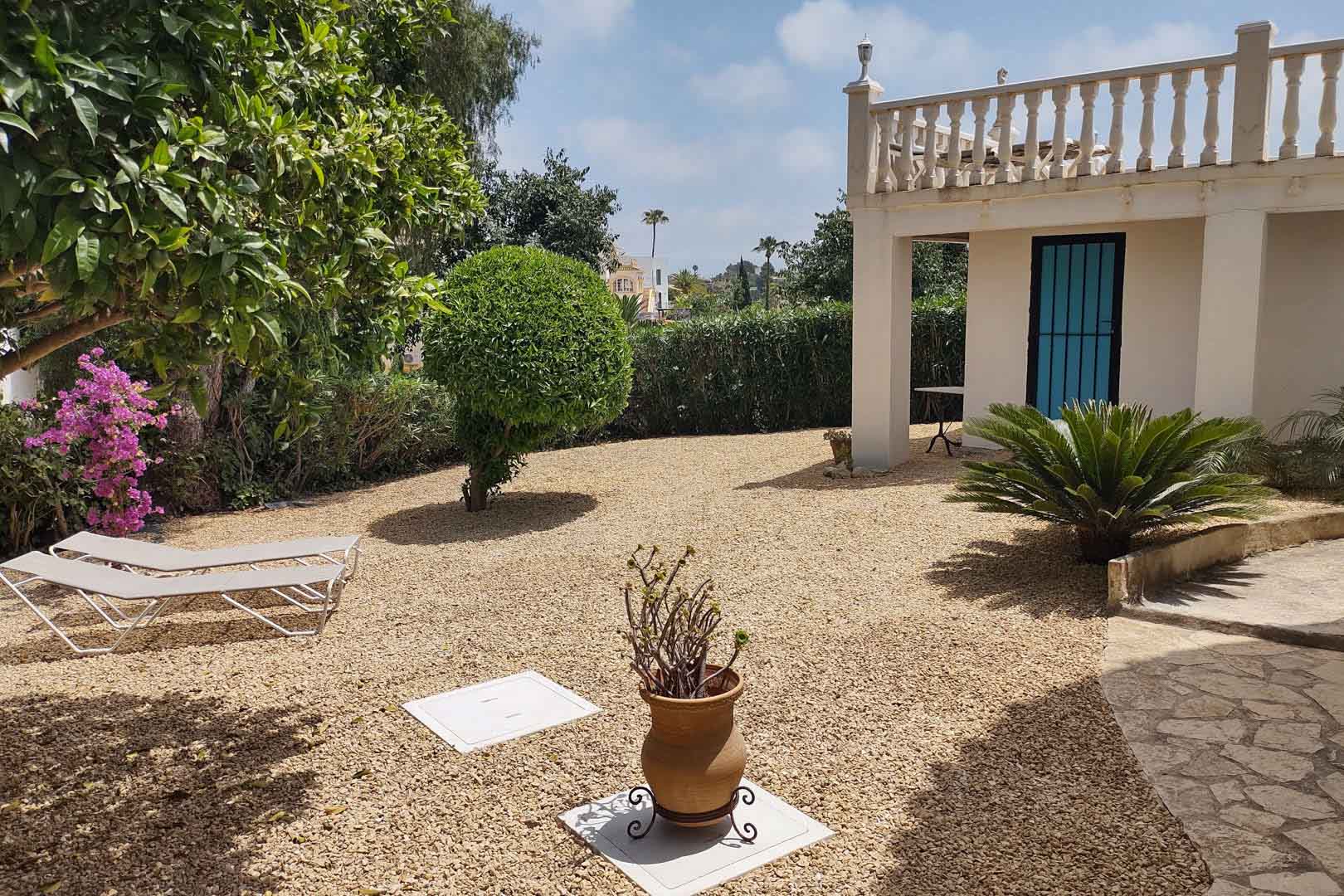 Jardin méditerranéen en gravier avec chaises longues, plantes locales et façade blanche agrémentée d'une balustrade classique et d'une porte bleue.