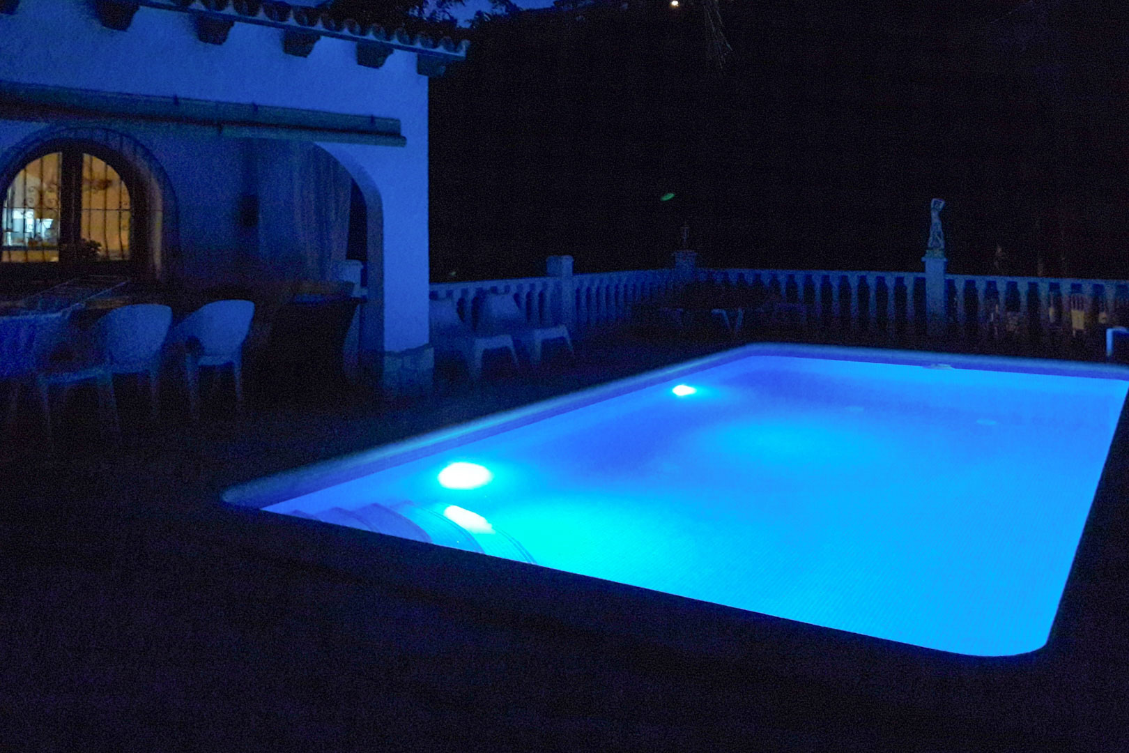 Piscine rectangulaire avec éclairage LED bleu nocturne, située à côté d'une terrasse couverte avec arches et une balustrade blanche traditionnelle.