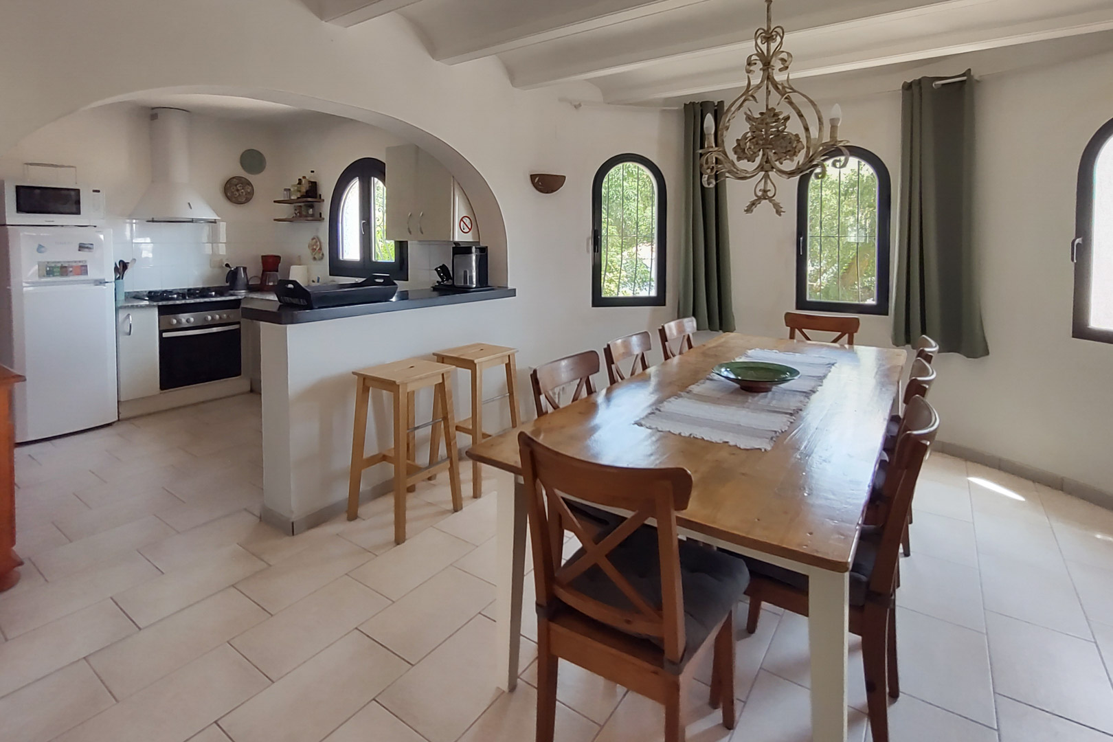 Salle à manger avec table en bois pour huit personnes, ouverte sur une cuisine équipée via une arche, comprenant un bar avec tabourets et des fenêtres cintrées.