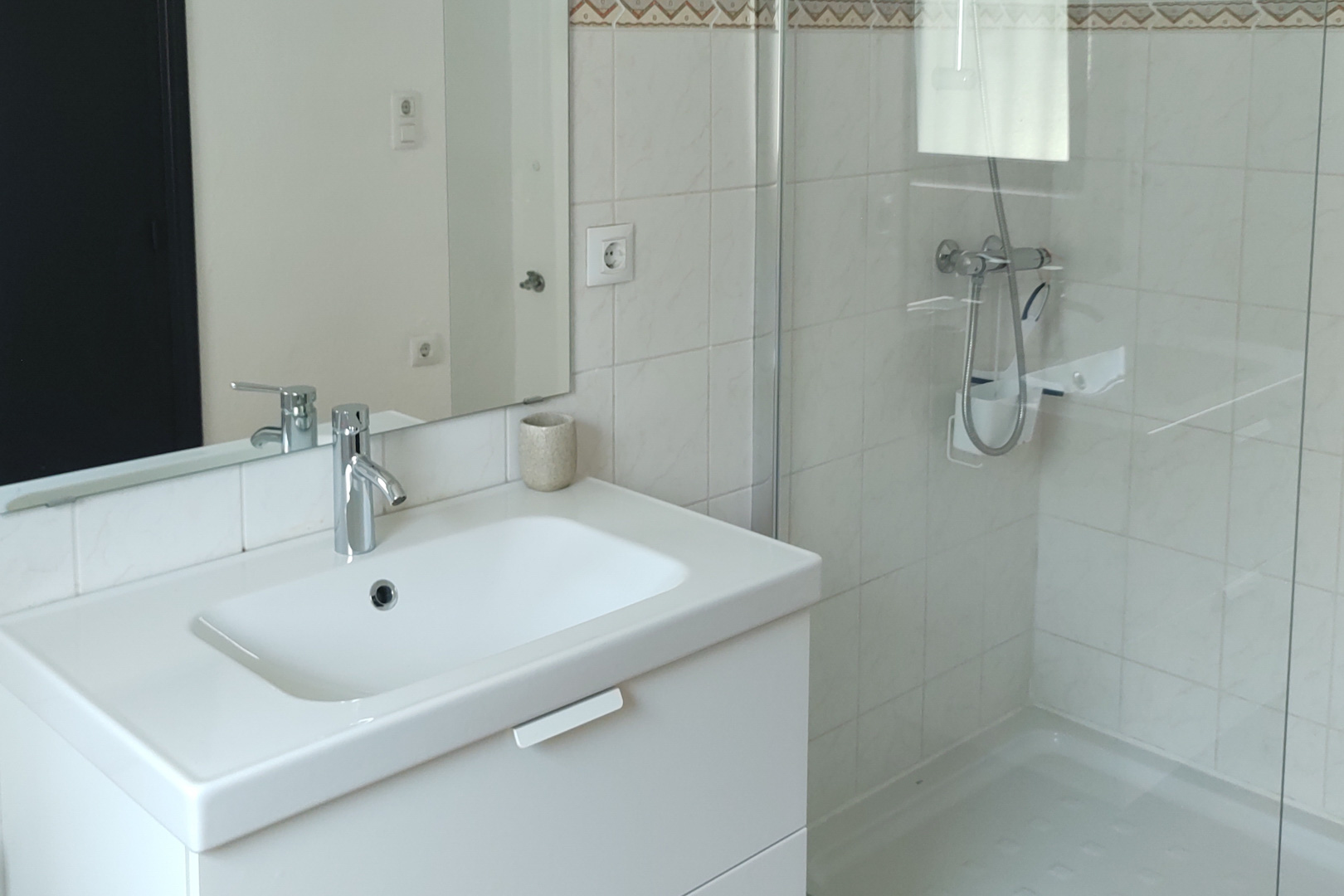 Salle de bain fonctionnelle avec meuble vasque blanc, robinetterie chromée et douche à l'italienne avec paroi vitrée. Murs carrelés en blanc avec frise décorative.